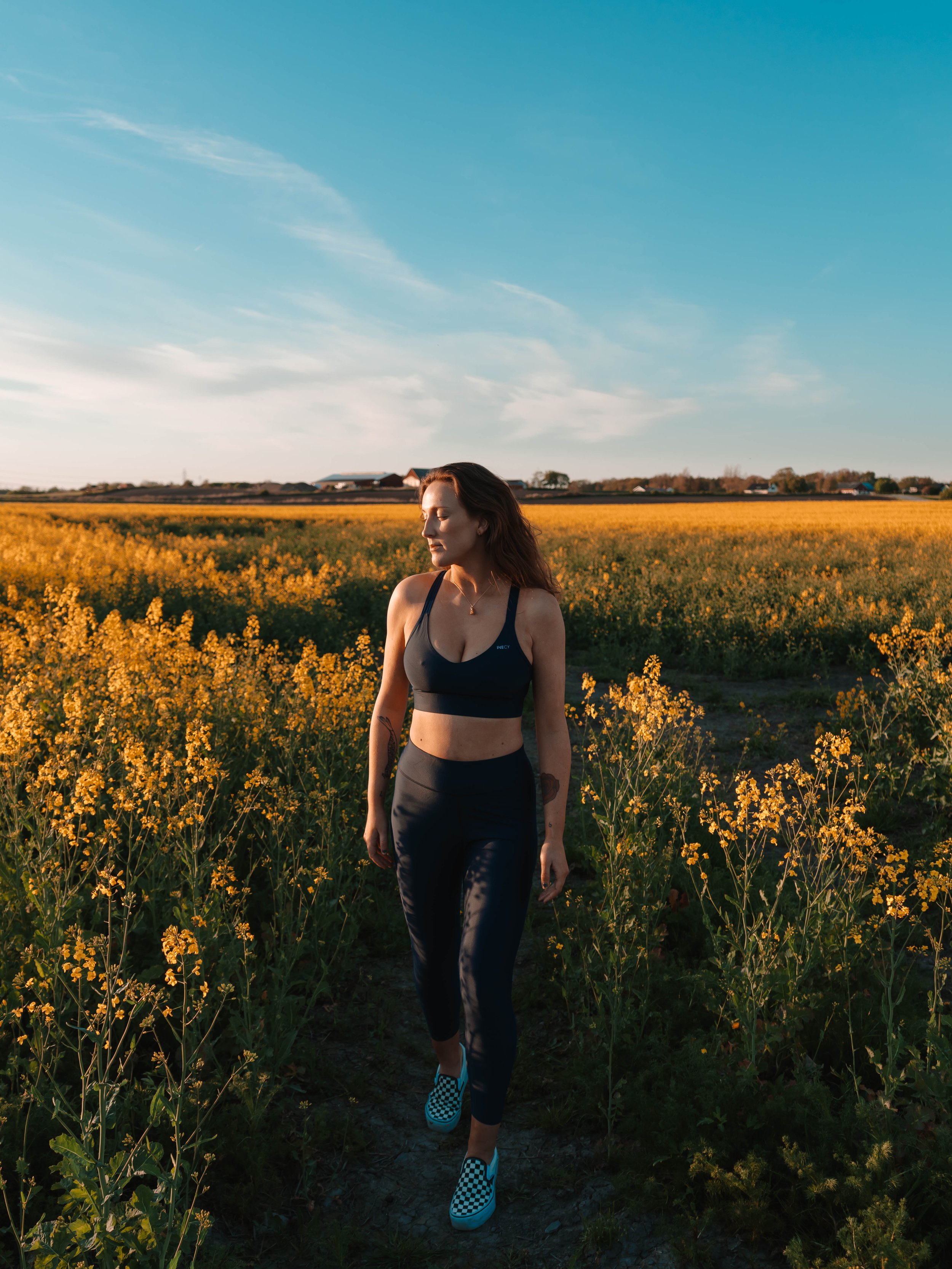 A woman walking along a narrow dirt path through a field of yellow flowers under a blue sky.