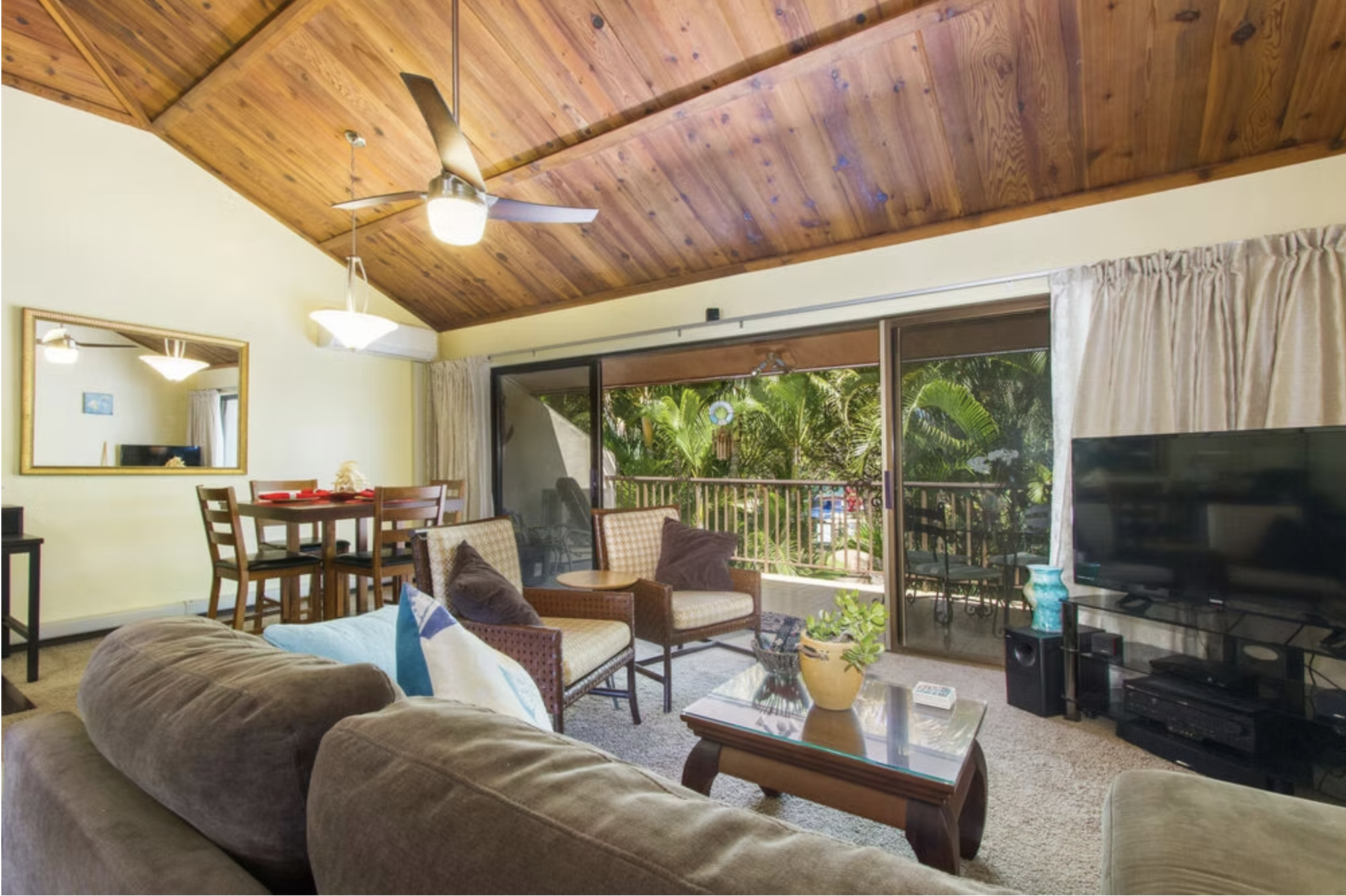 Living room with a beige sofa, glass coffee table, armchairs, large flat-screen TV, and a sliding glass door leading to a balcony with tropical plants.
