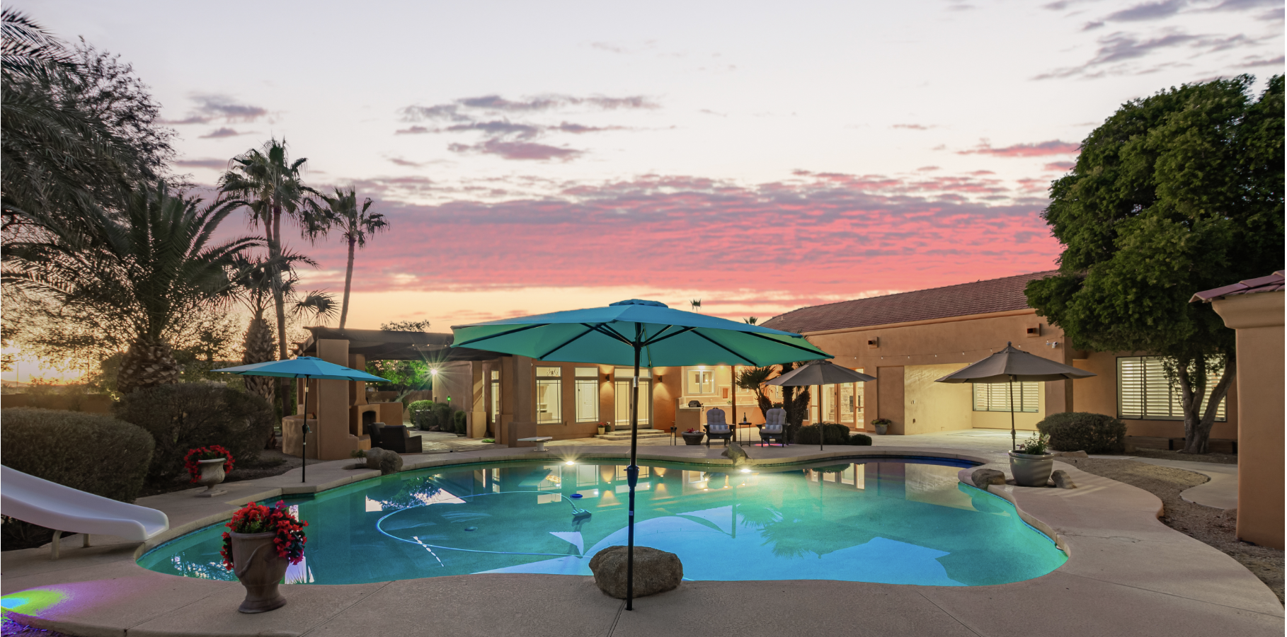 A backyard swimming pool area during sunset with pink and orange sky. The pool has blue lighting, and there are blue and beige umbrellas with outdoor chairs around. There are potted plants, bushes, a large tree, and a house with lit windows in the background.