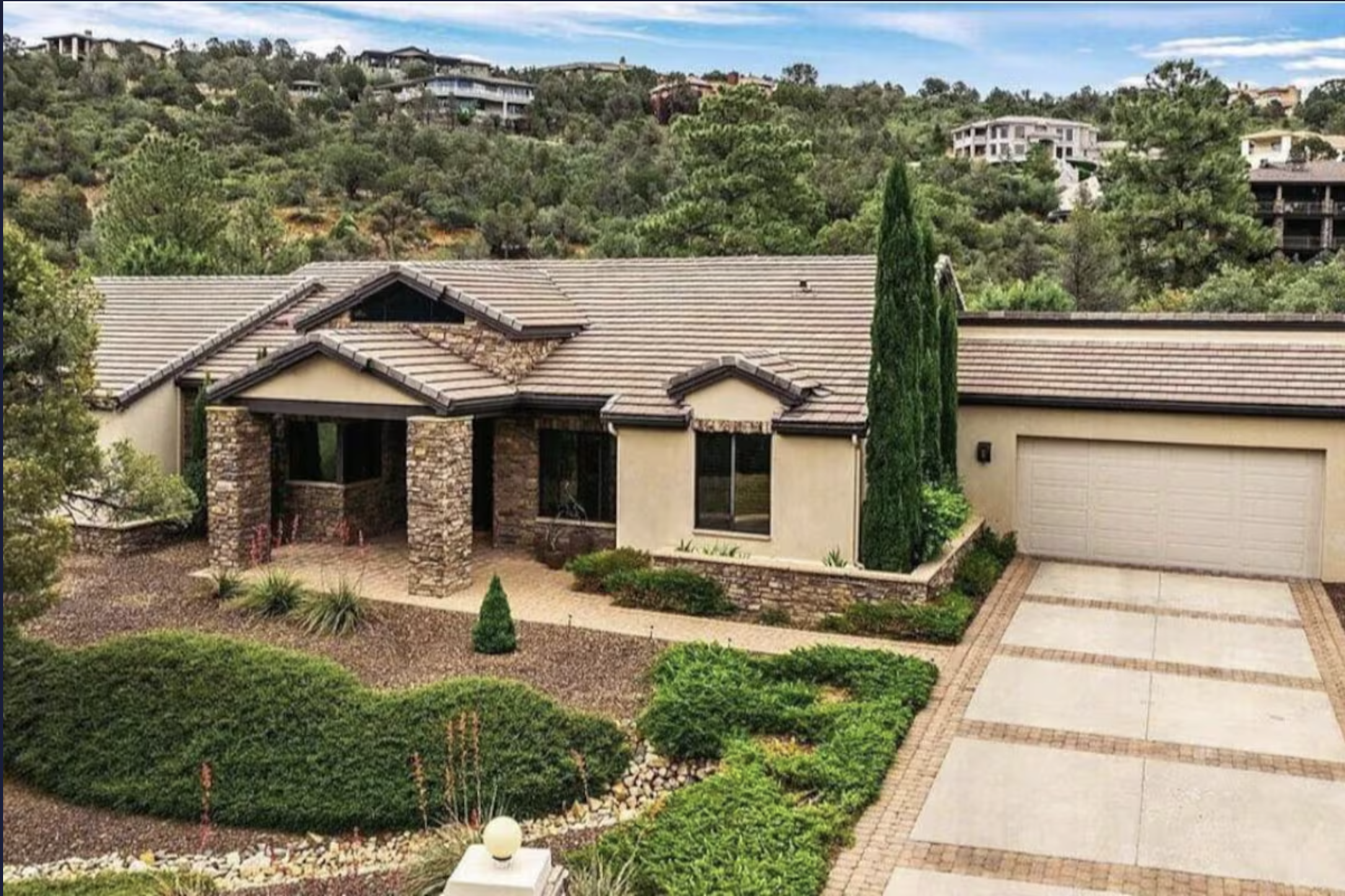 Front yard of a house with a paved walkway, landscaped garden beds, small shrubs, and a large cypress tree.