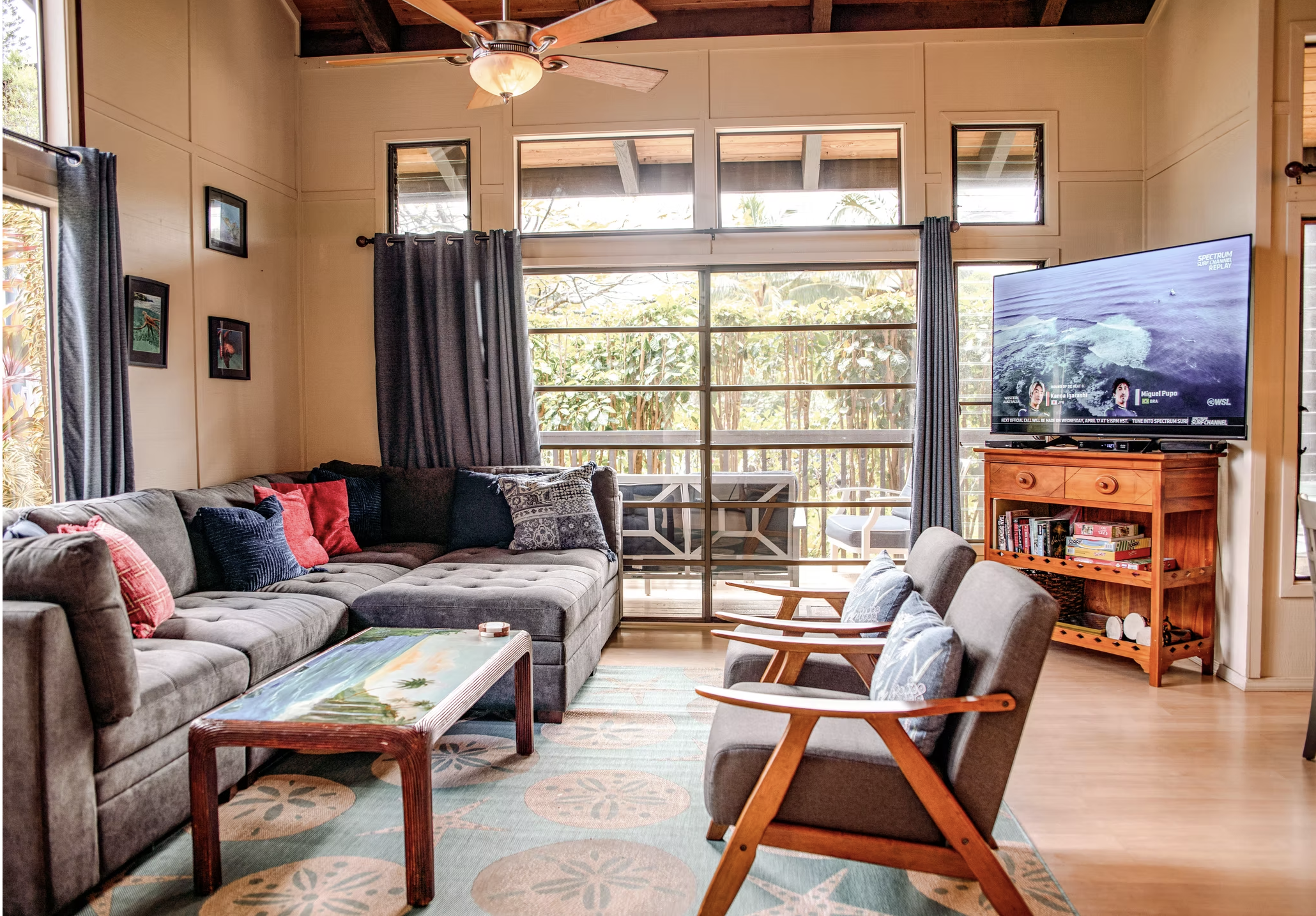 Living room with large windows, a gray sectional sofa with colorful pillows, a coffee table with a tropical image, a wooden rocking chair, a TV on a wooden stand, and framed photos on the wall. Outdoors greenery is visible through the windows.