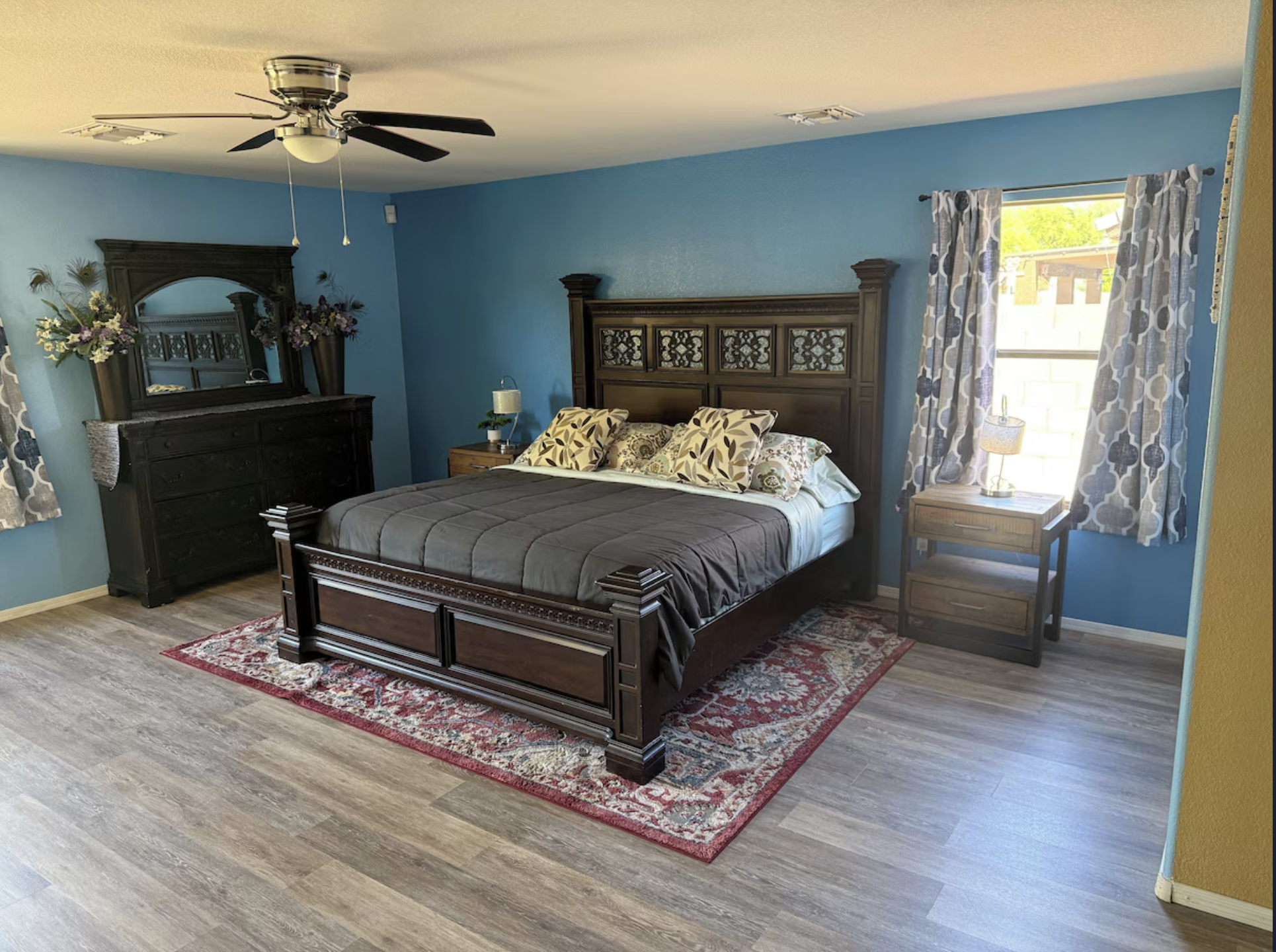 A bedroom with blue walls, a dark wooden bed with a headboard, and matching dresser with a mirror. The bed has patterned pillows and a comforter. There are side tables with lamps, curtains with a geometric pattern, a ceiling fan, and a window showing outside greenery.
