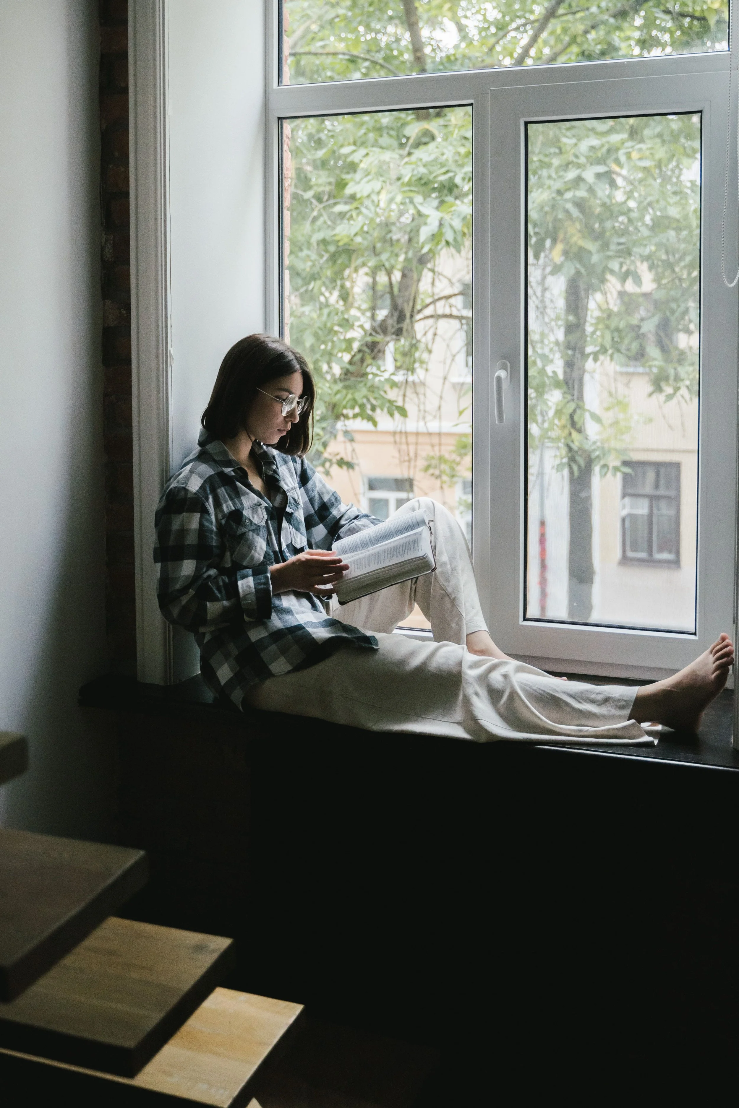Woman sitting by a window reading quietly, reflecting on questions of faith and identity