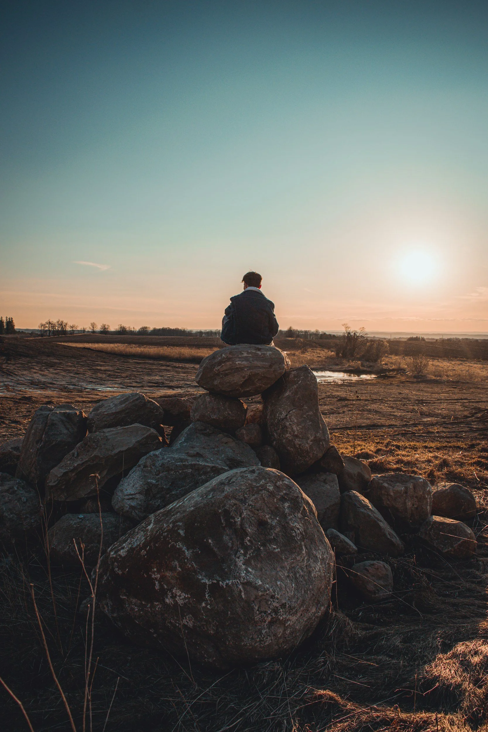 Person sitting alone on rocks watching the sunset, representing quiet contemplation on faith and identity