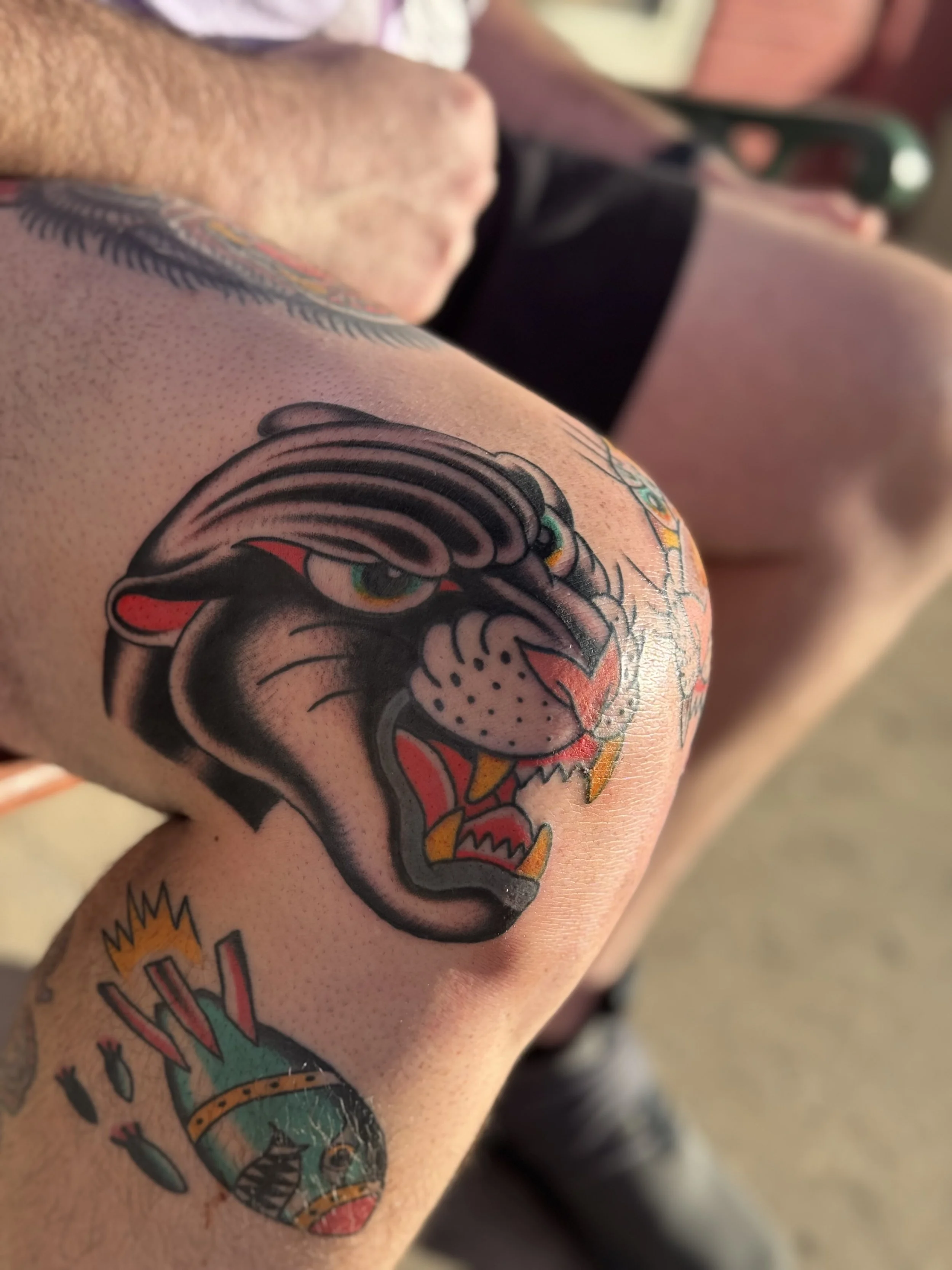 Close-up of a person's upper arm with a colorful tattoo of a fierce animal, possibly a tiger or a dragon, with vivid red, black, and green colors.