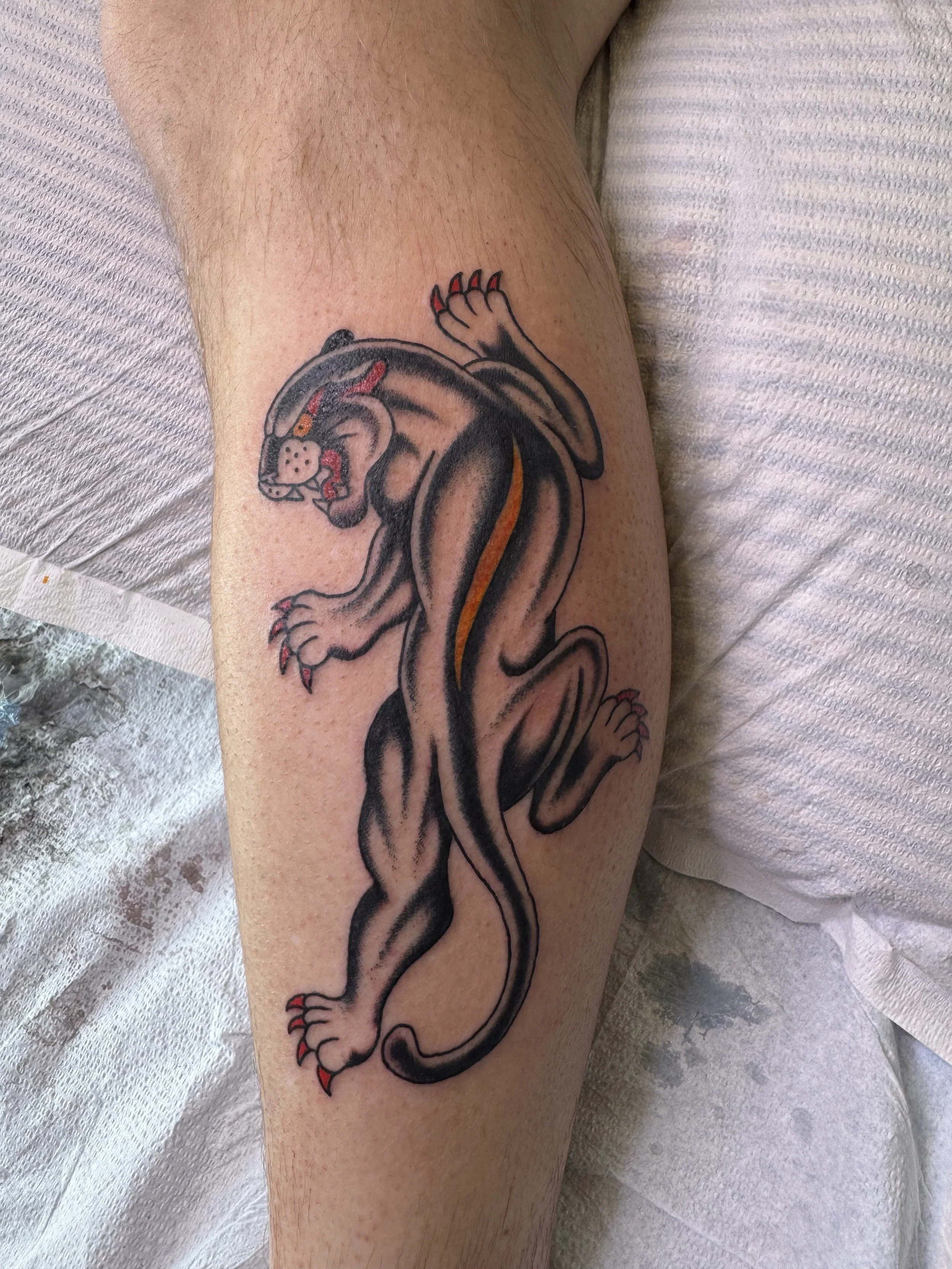 Tattoo of a panther on a person's leg, with the panther depicted in black and orange with red details on its claws and tongue.