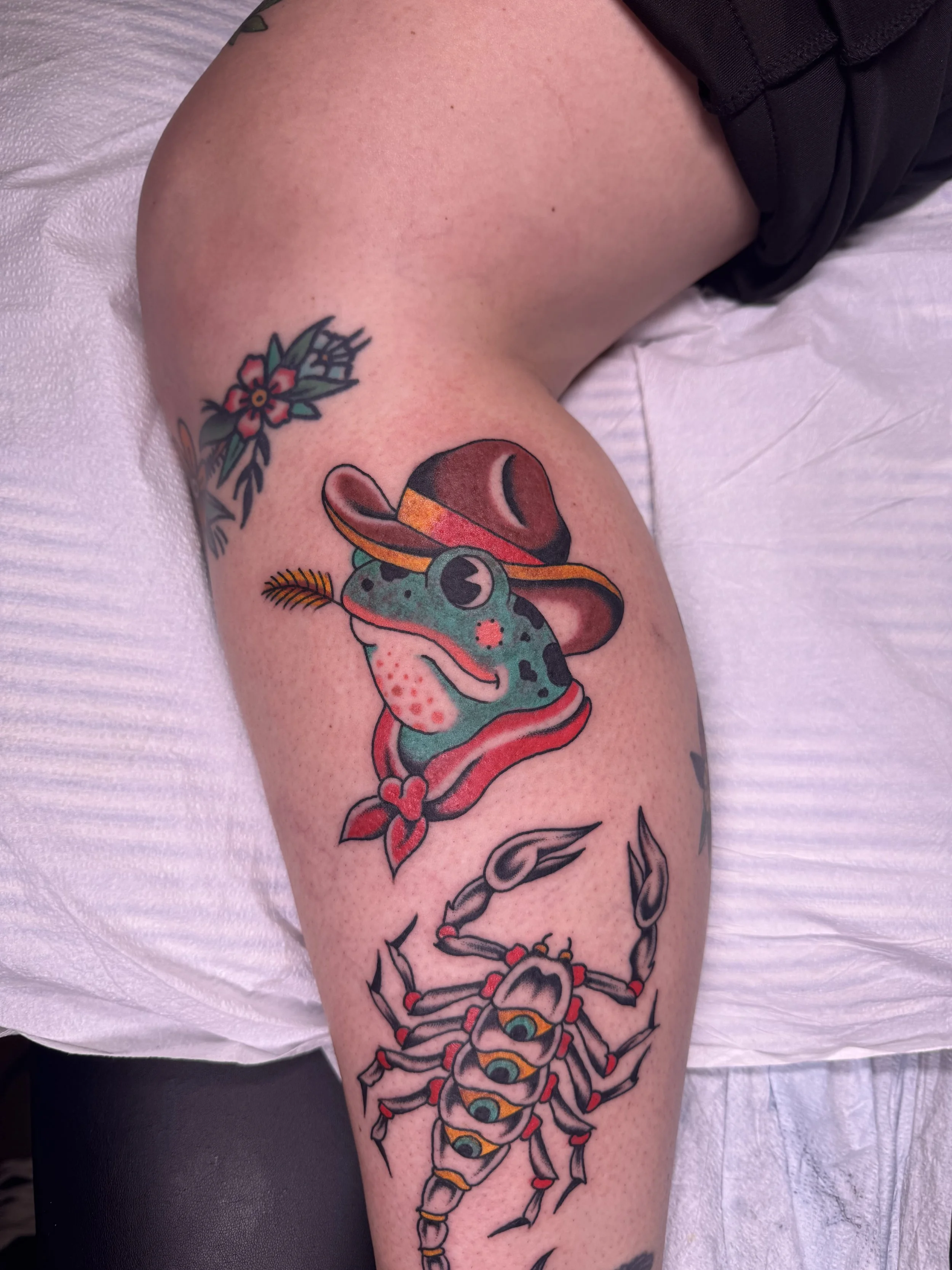 Tattoo of a gecko with colorful eyes on a person's thigh, surrounded by additional tattoo designs including a spider and floral elements.