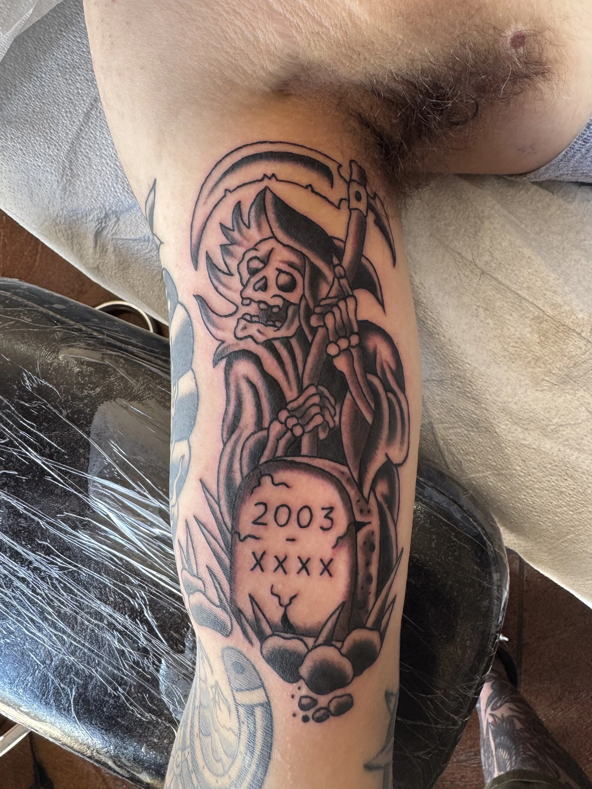 Tattoo of a skeleton with a scythe, a rainbow, and a skull inscribed with the year 2003 and five X's on the forearm.