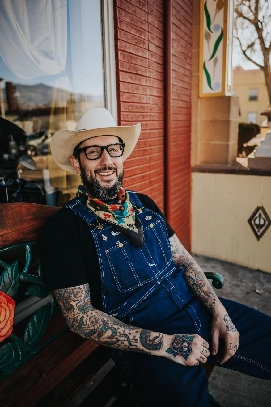 A smiling man with a beard and tattoos, wearing a cowboy hat, glasses, a bandana, and overalls, sitting on a park bench outside a building with red brick walls.