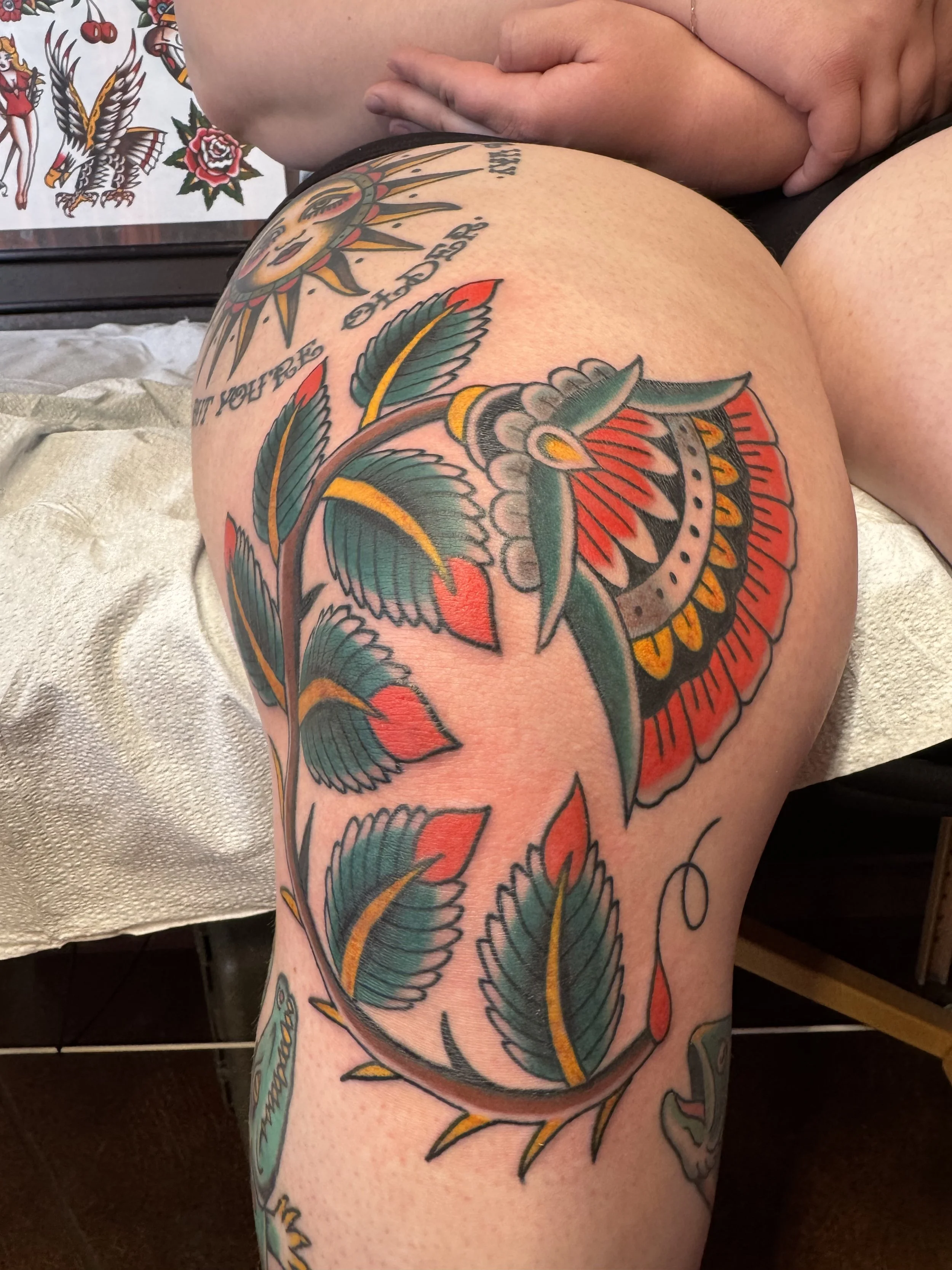 Close-up of a person's thigh and hip, showcasing a colorful tattoo featuring feathers, leaves, and floral designs.