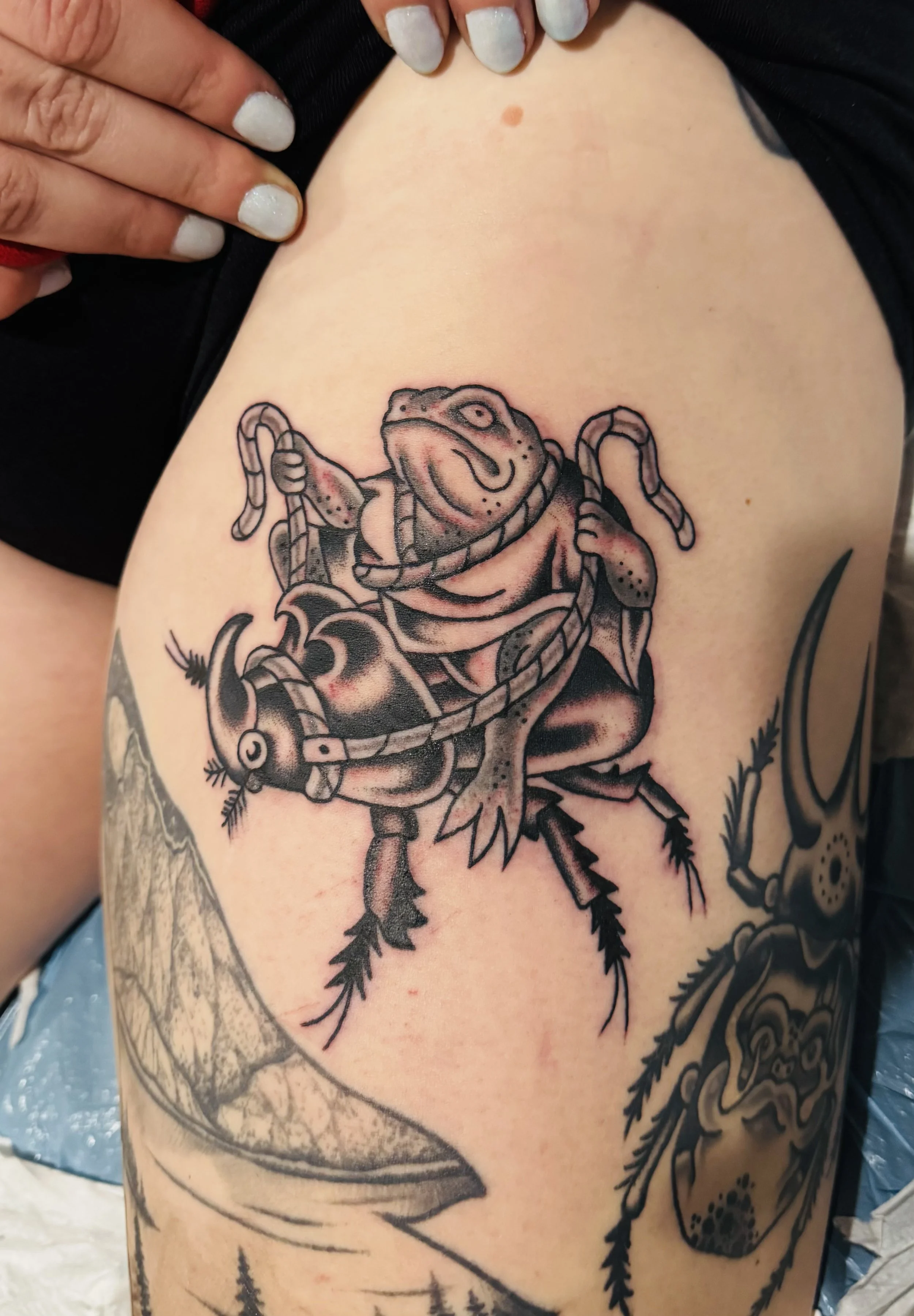 A person with a tattoo of a frog holding a snake on their upper arm and side, with other tattoos visible.