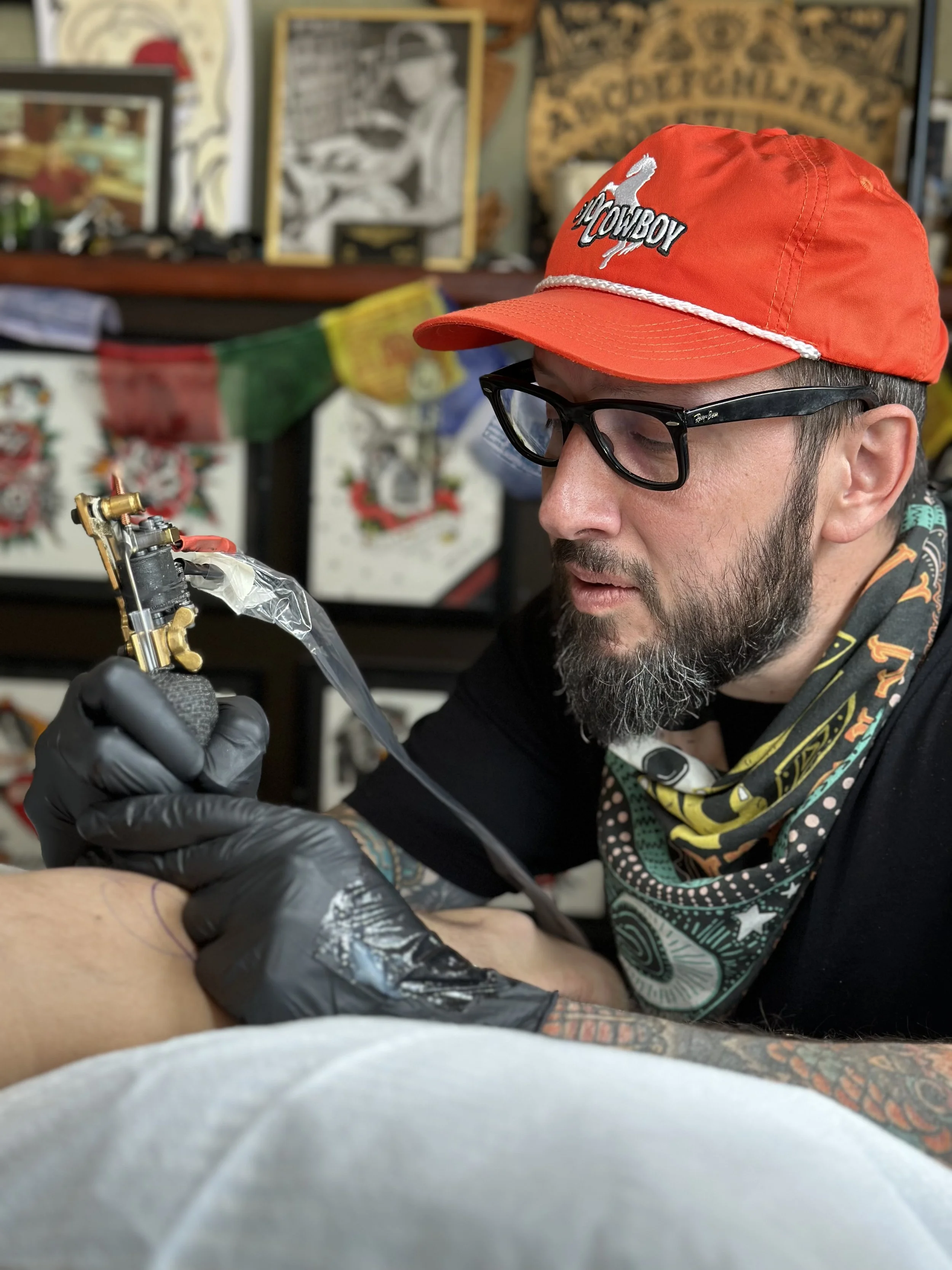 A man with a beard, glasses, and a red cap labeled 'Cowboy' is tattooing someone's arm in a tattoo studio with various colorful artwork and decorations in the background.