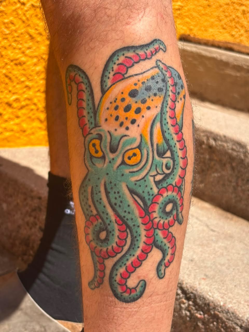 Colorful tattoo of an octopus with yellow and black eyes, greenish skin, and red and pink tentacles, on a person's leg.