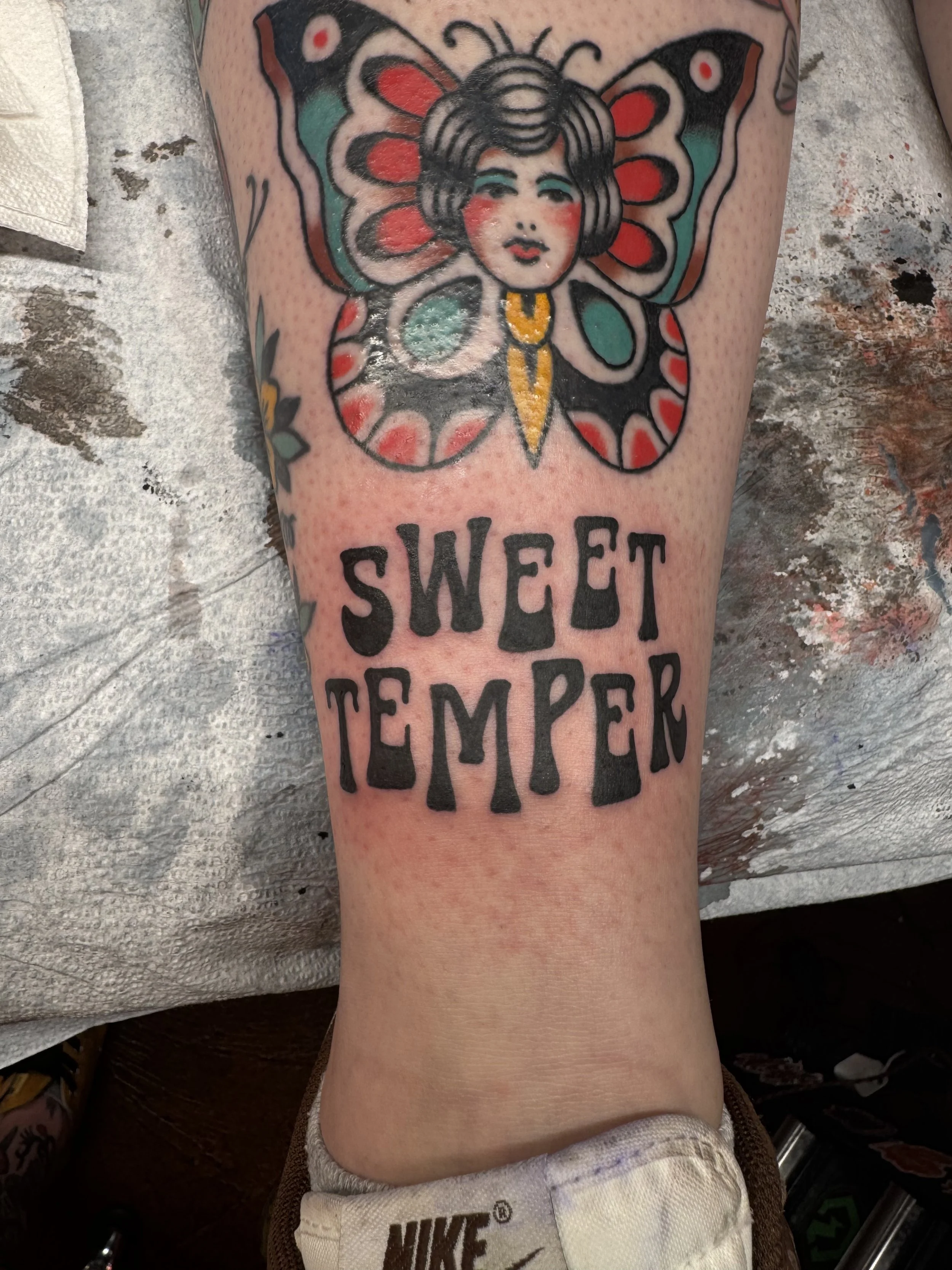 Tattoo of a butterfly with a woman's face in the center, featuring black hair, blue eyeshadow, and red cheeks. The butterfly's wings are decorated with red, black, and turquoise patterns. Below the butterfly, the words "SWEET TEMPER" are inked in bold black letters.
