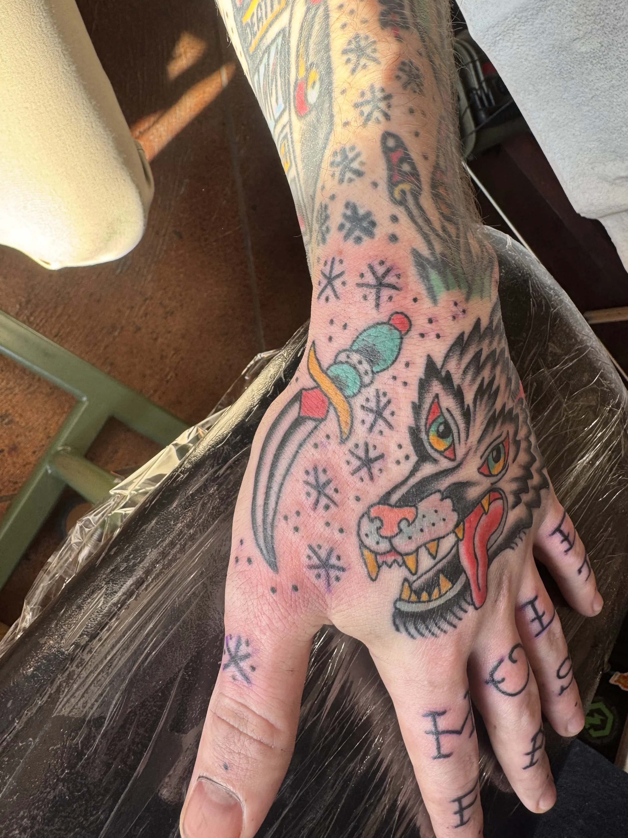 A tattooed hand with colorful ink, featuring abstract and cartoonish designs, including a large wolf head, snowflakes, a snake, and various geometric patterns.