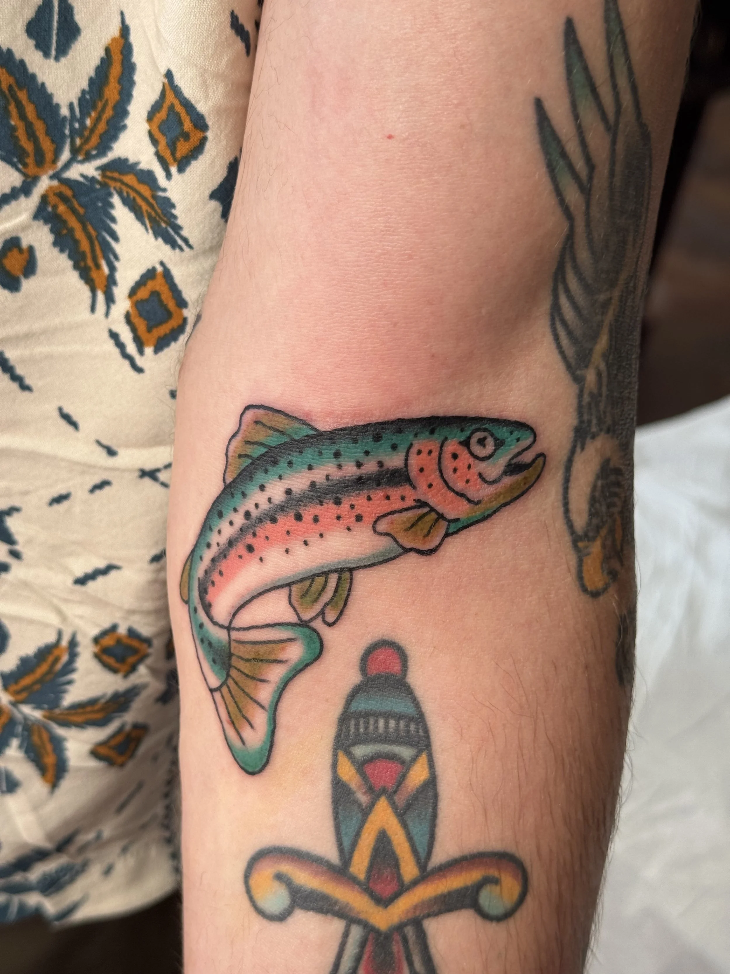 A person's arm with colorful tattoos of a rainbow trout fish and geometric designs, alongside a patterned fabric with orange and blue designs.