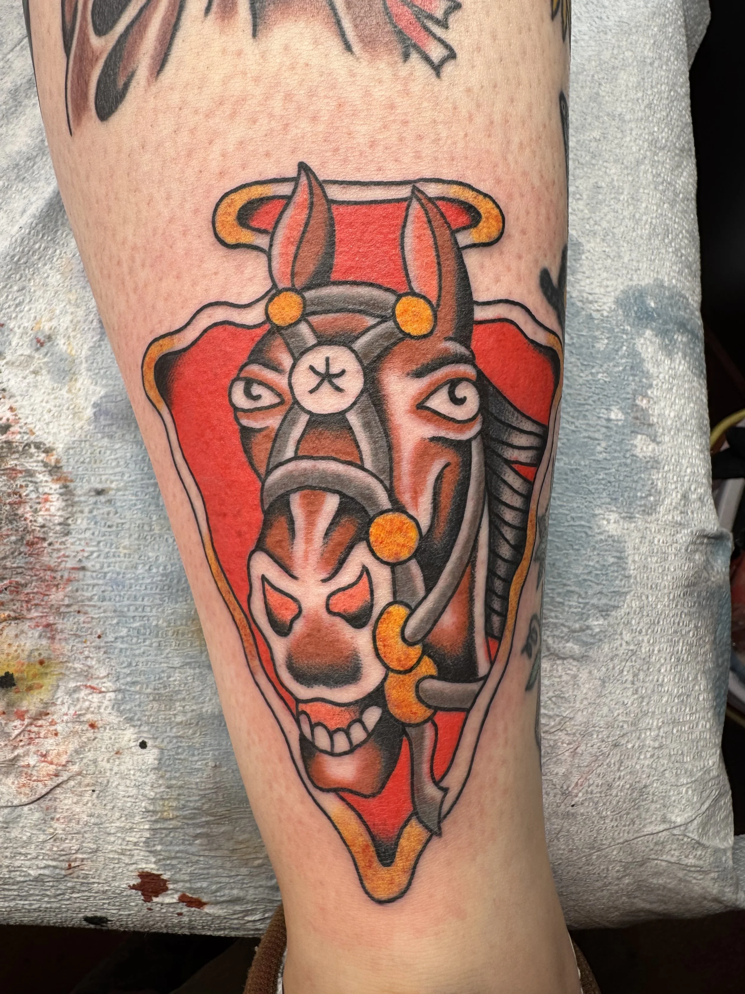 Tattoo of a horse with a stylized, fierce expression, featuring red, black, and orange colors, on a person's upper arm.