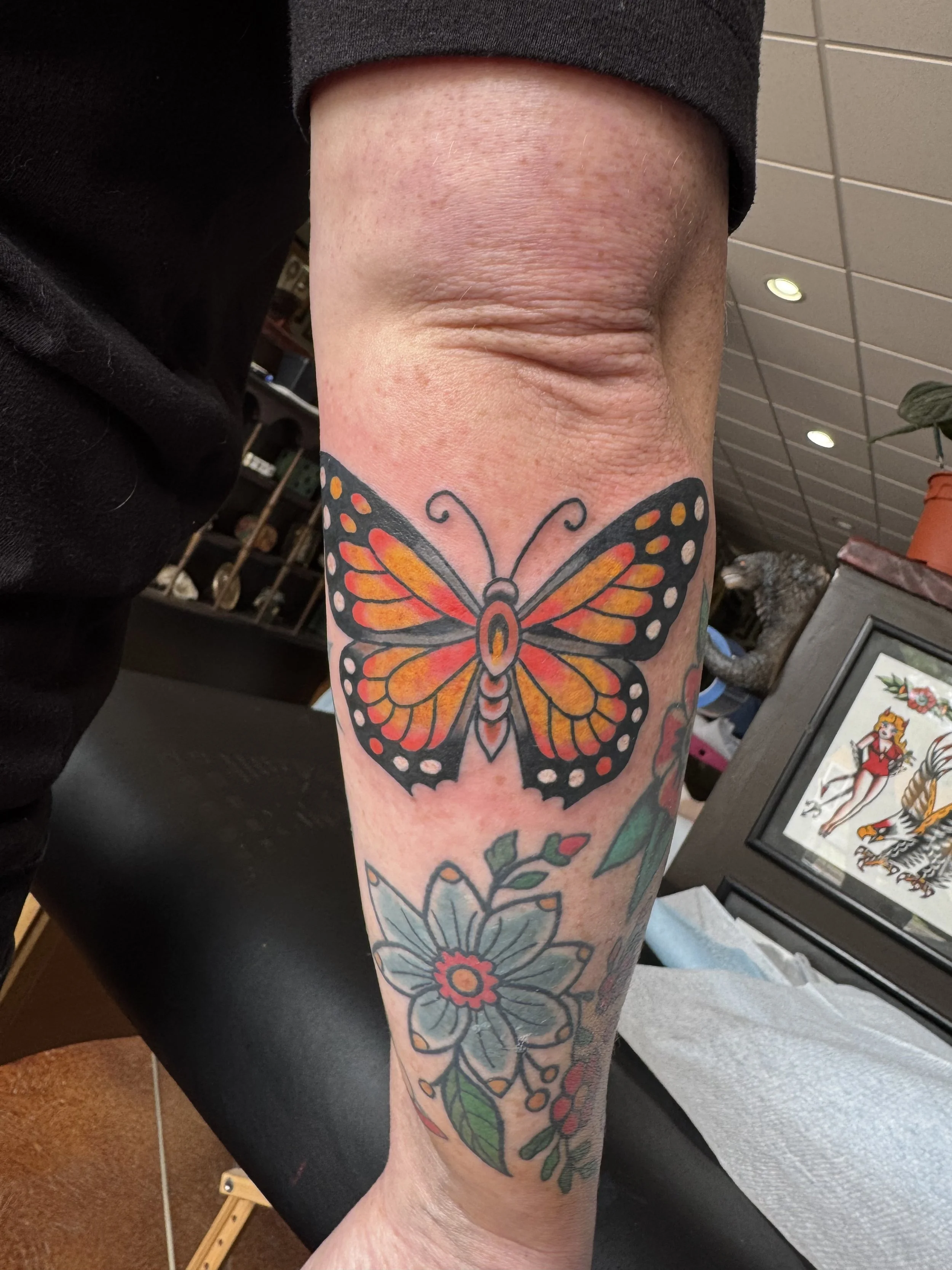 A person's forearm with a colorful butterfly tattoo, featuring orange, black, and white wings, as well as a floral design below.