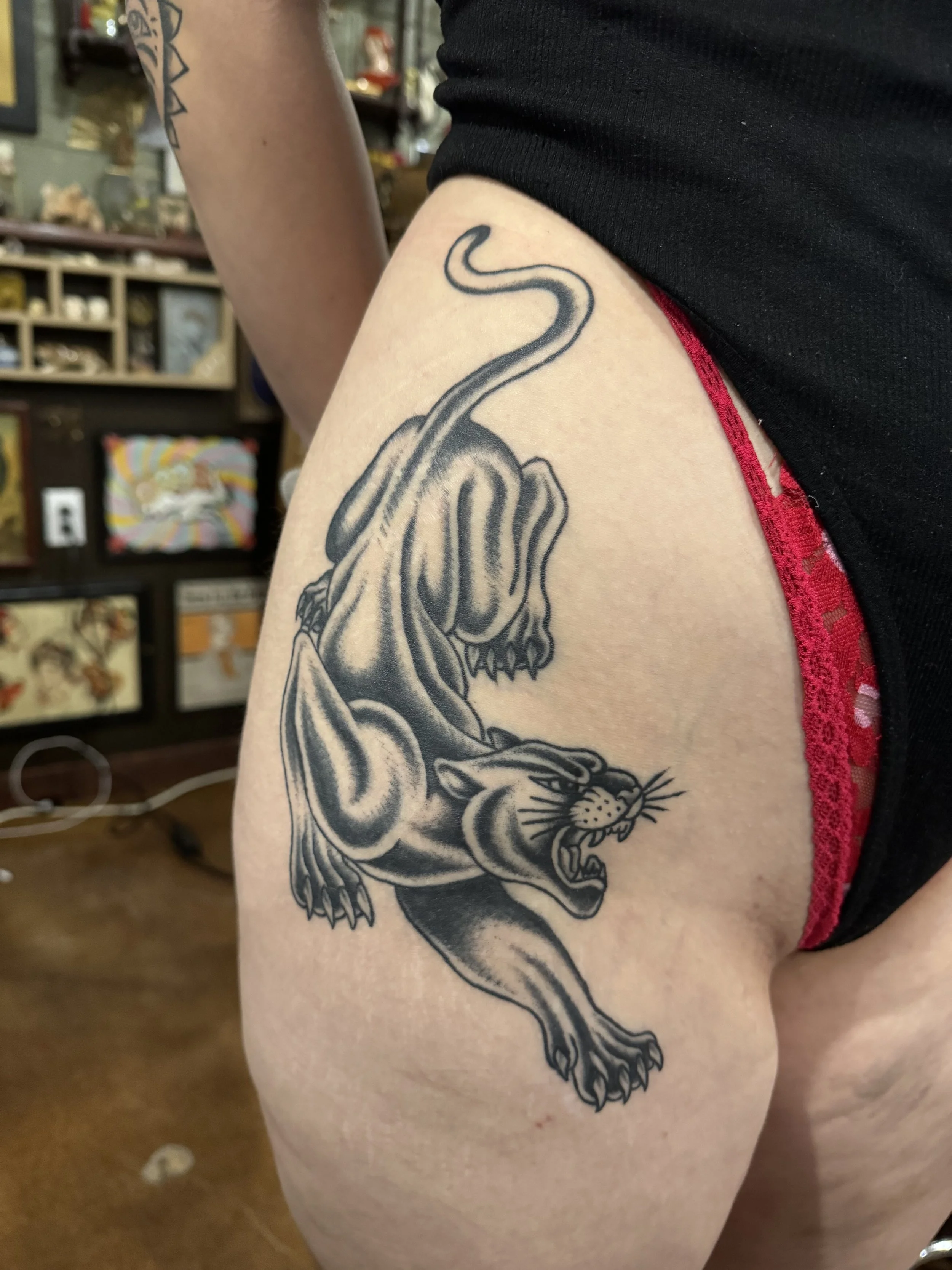 Tattoo of a fierce lion's head on a person's upper thigh, with the lion's mouth open revealing sharp teeth, detailed mane, and wild expression, with some text partially visible.