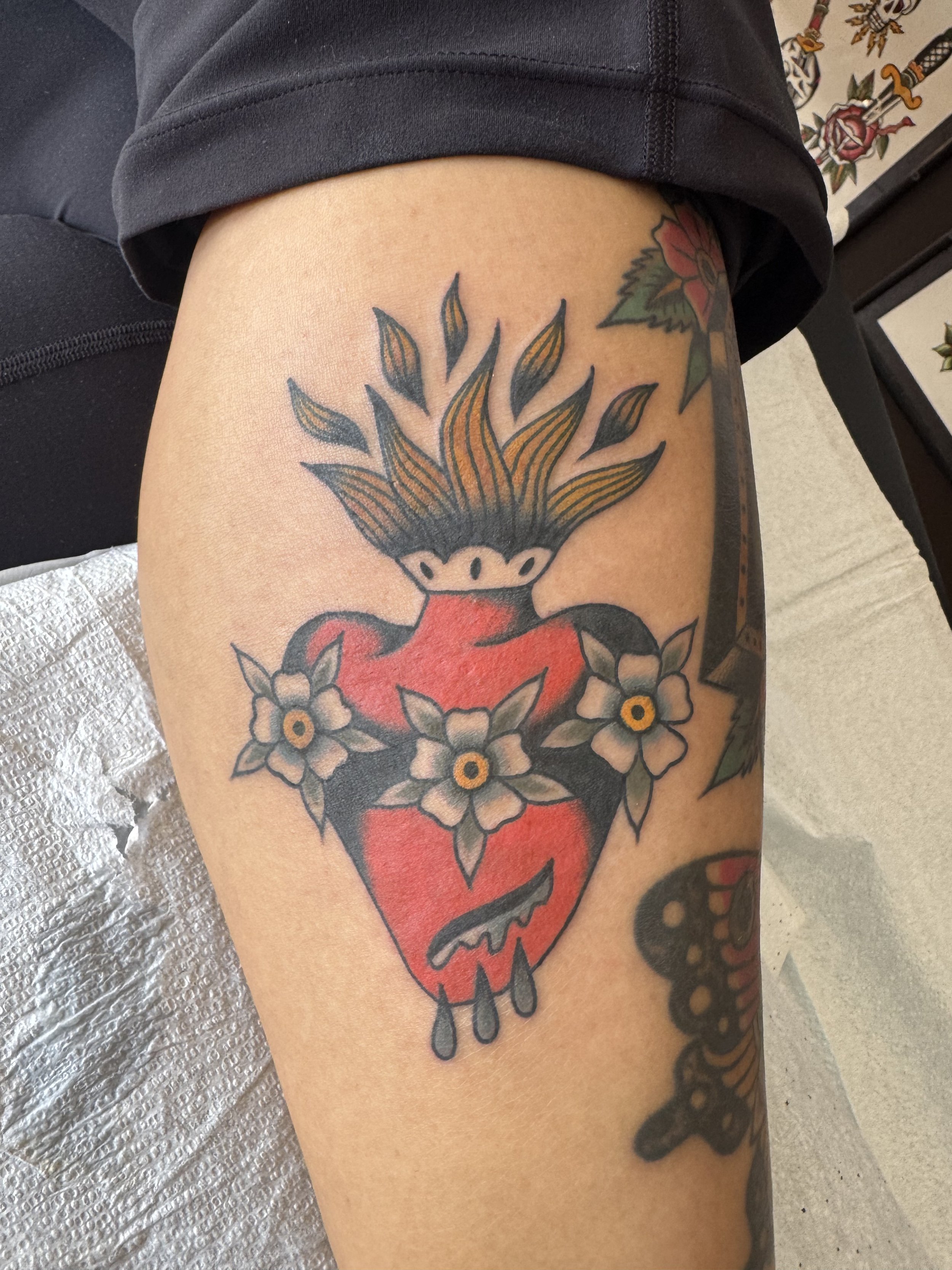 A tattoo of a red heart with a spooky face, surrounded by white flowers with yellow centers and green leaves on a person's arm.