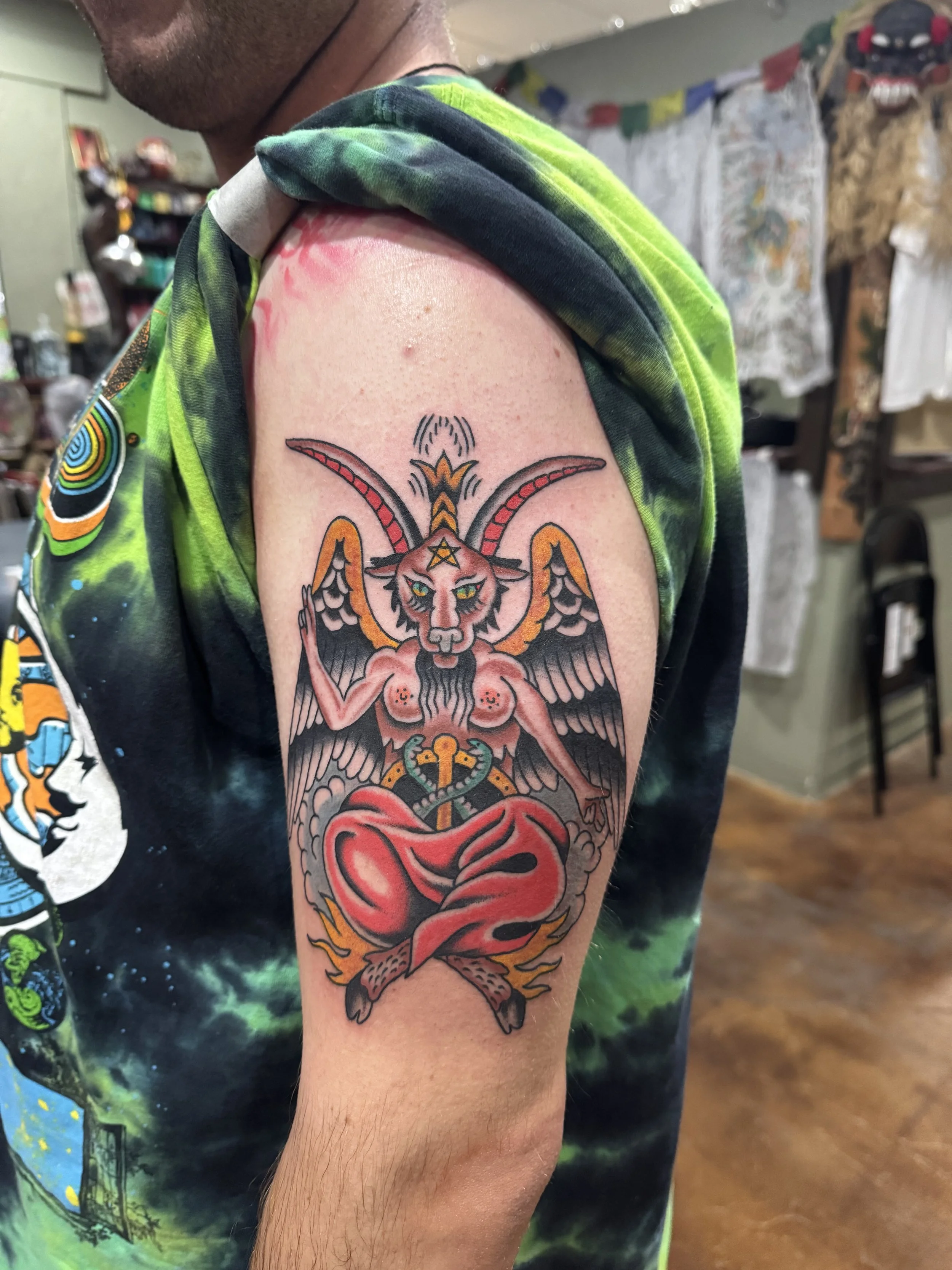 Tattoo of a goblin or demon face with horns, wings, and a snake below, on a person's upper arm. The individual is wearing a colorful tie-dye shirt and is in a room with various decorations in the background.