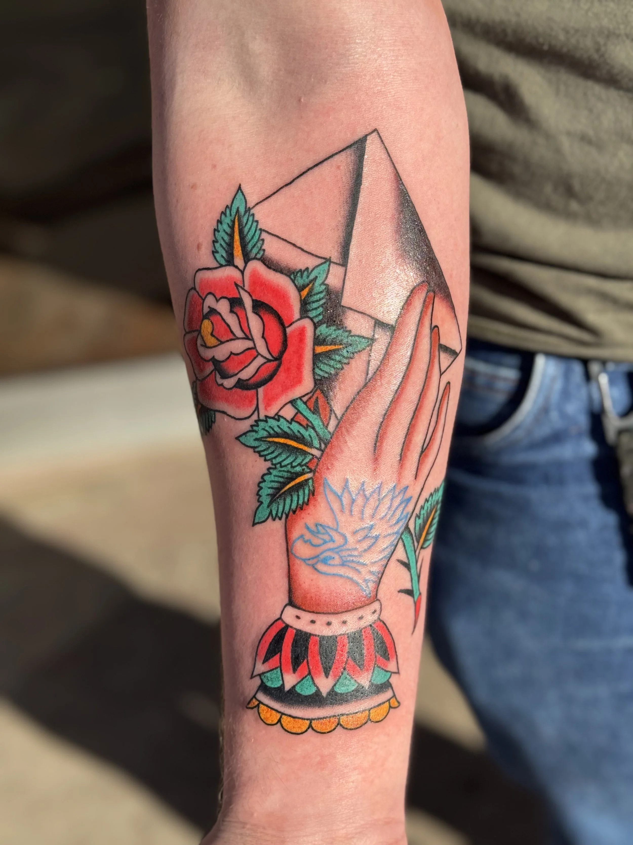 A colorful tattoo of a traditional-style woman with long hair, wearing a headdress, holding an urn with orange flowers at the top, and surrounded by vibrant flowers and leaves.