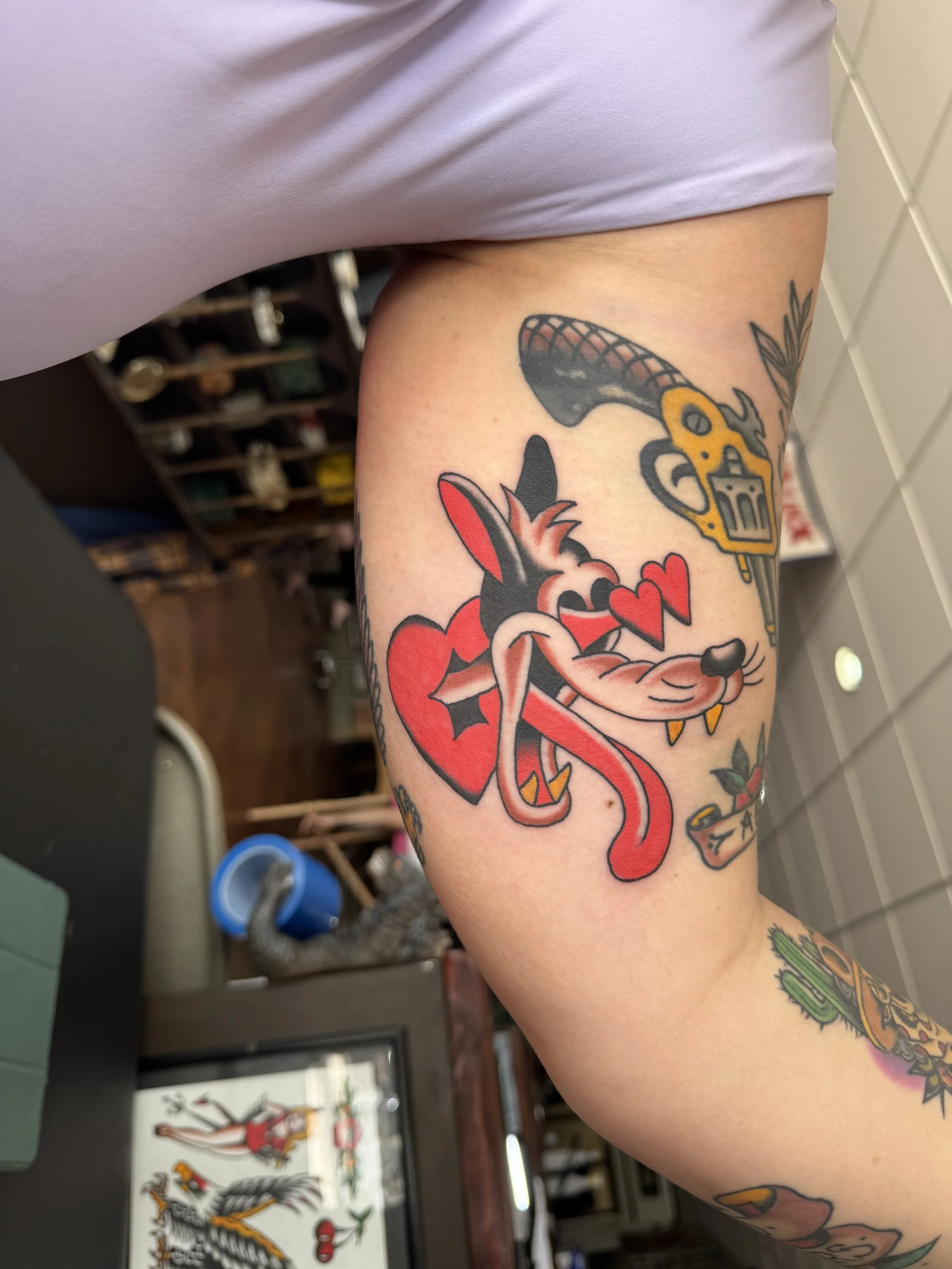 Close-up of a tattooed arm featuring a cartoonish snake intertwined with a heart, a dagger, and a sprig of leaves.