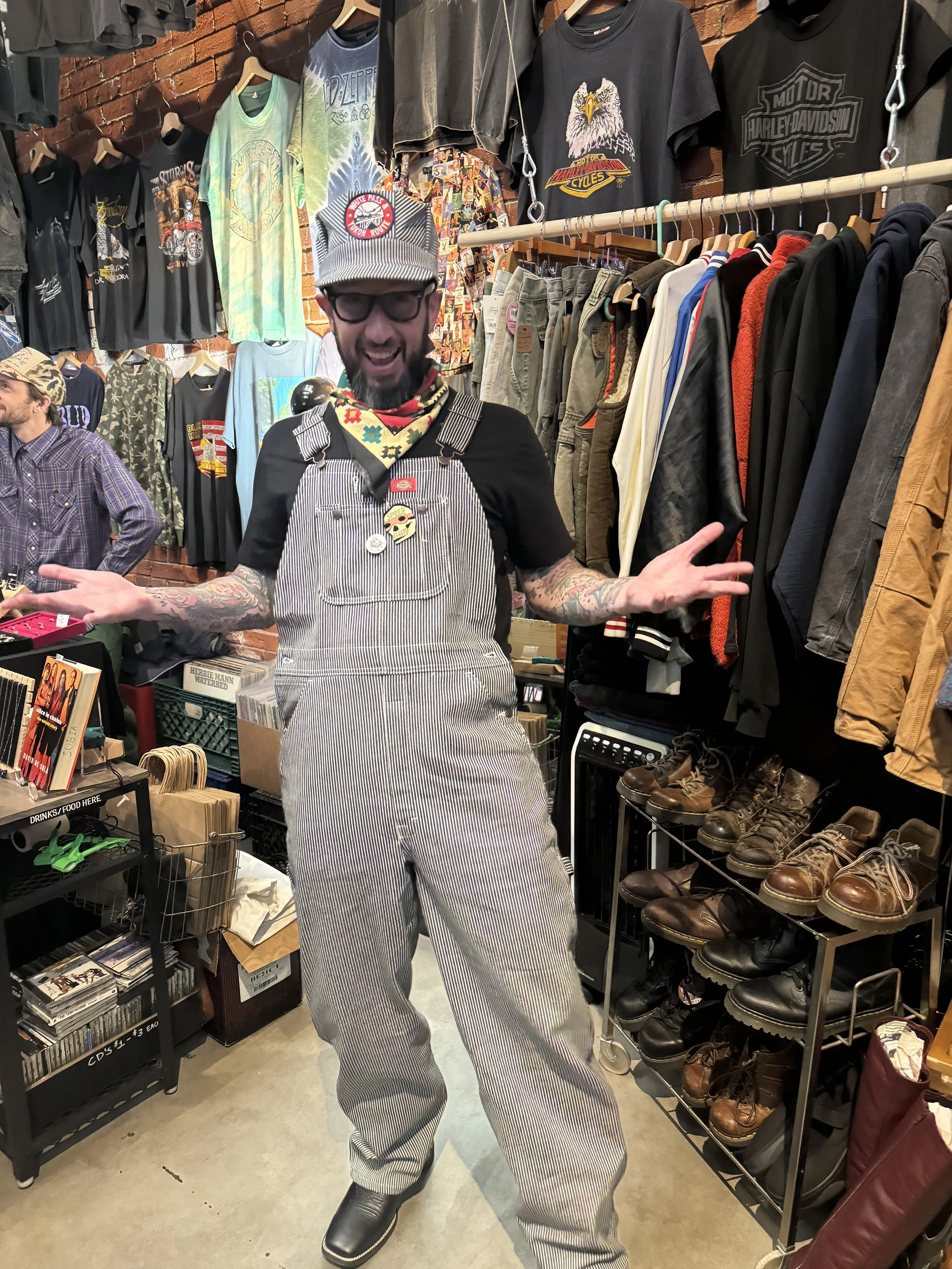 A man with glasses and a beard wearing a cap, bandana, black shirt, and striped overalls standing inside a clothing store with brick walls, surrounded by racks of T-shirts, pants, and shoes.