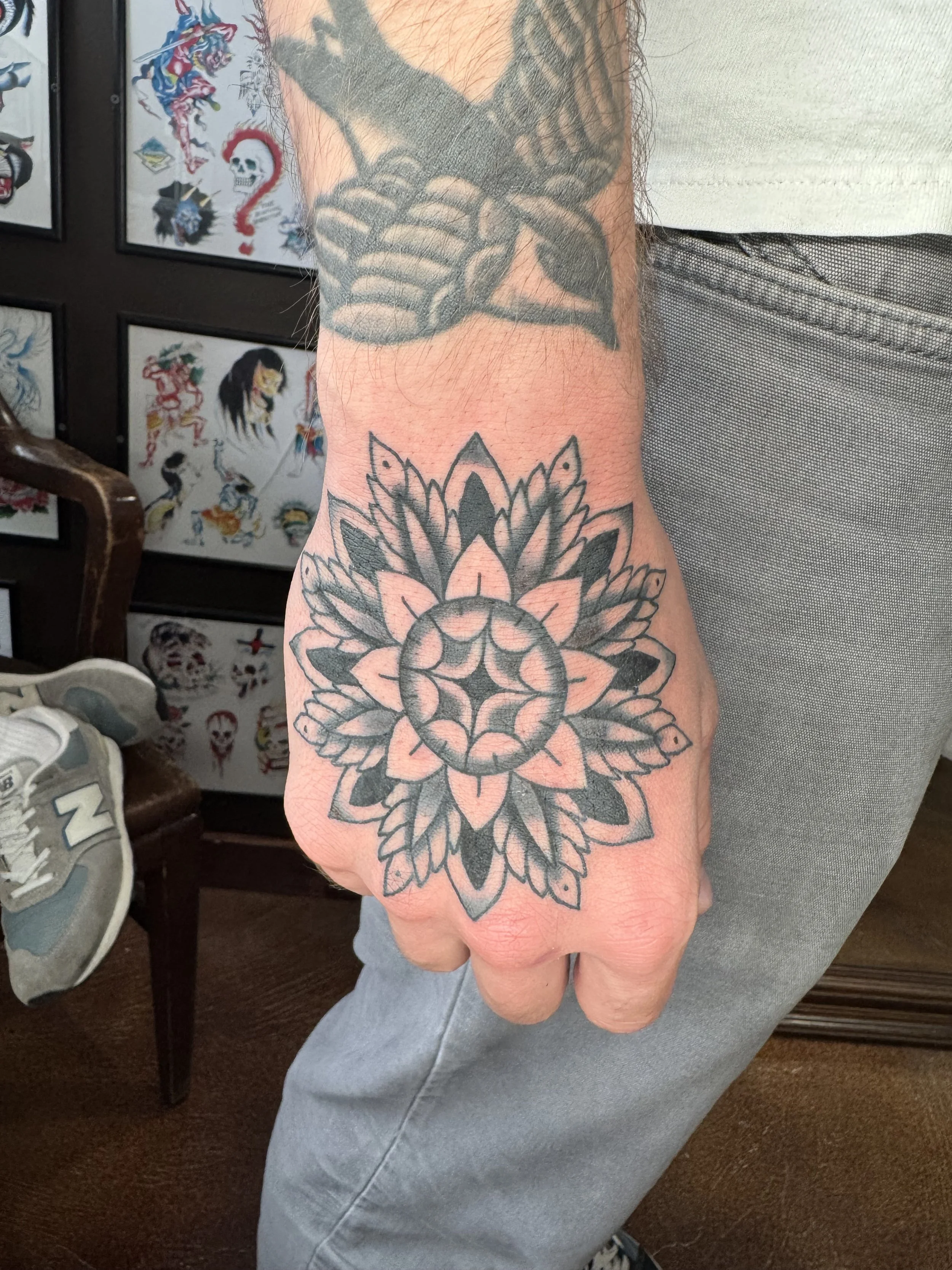 Tattoo of a mandala design on the forearm, featuring intricate black outline patterns. In the background, tattoo flash art is visible on the wall, including traditional American-style tattoo designs like skulls, animals, and other motifs.