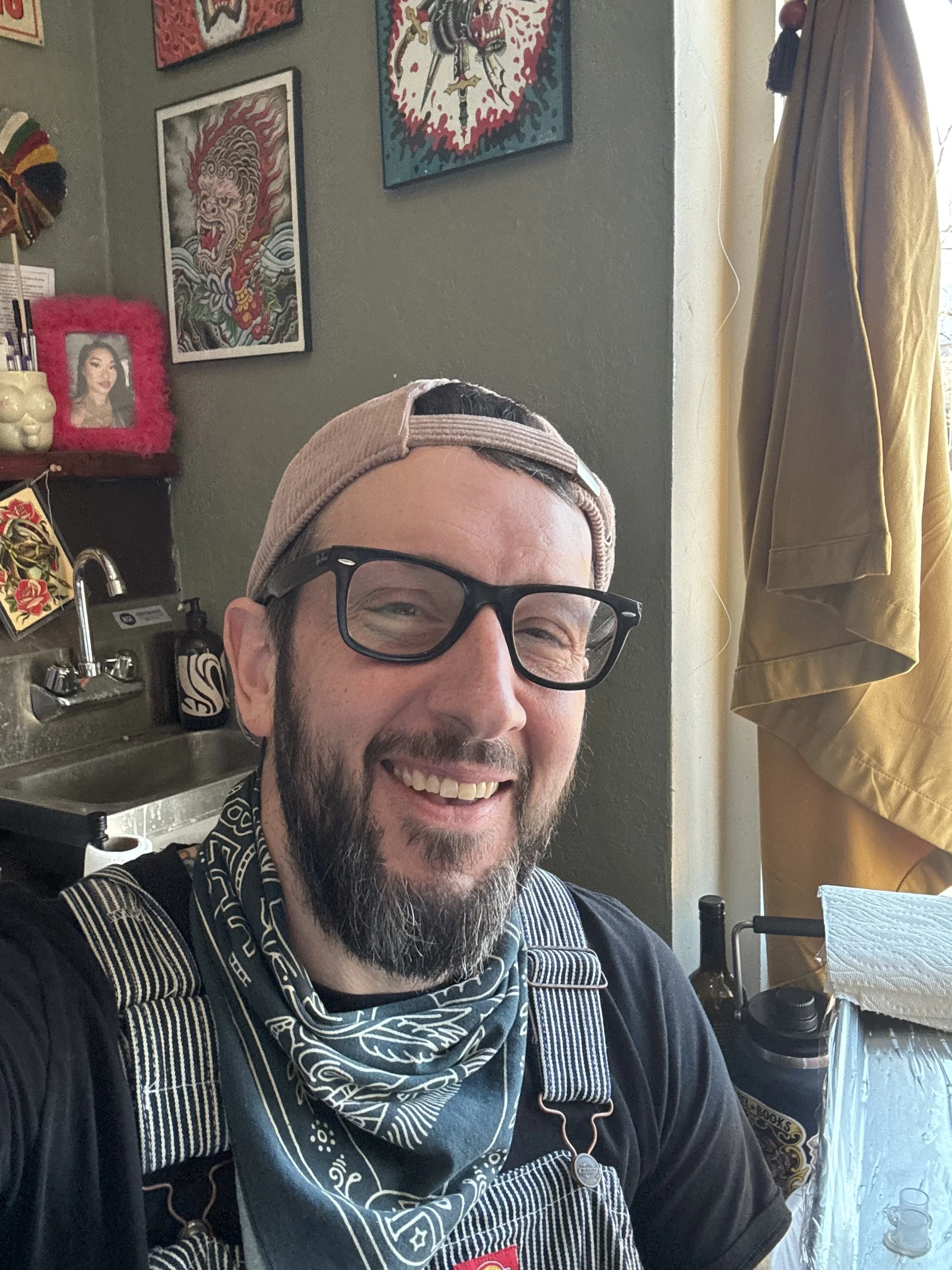 A man smiling in an american traditional tattoo shop, wearing a beige baseball cap backward, glasses, a black shirt, and a patterned scarf. Behind him is a green wall with framed artwork and a small photograph of a woman in a pink fuzzy frame.
