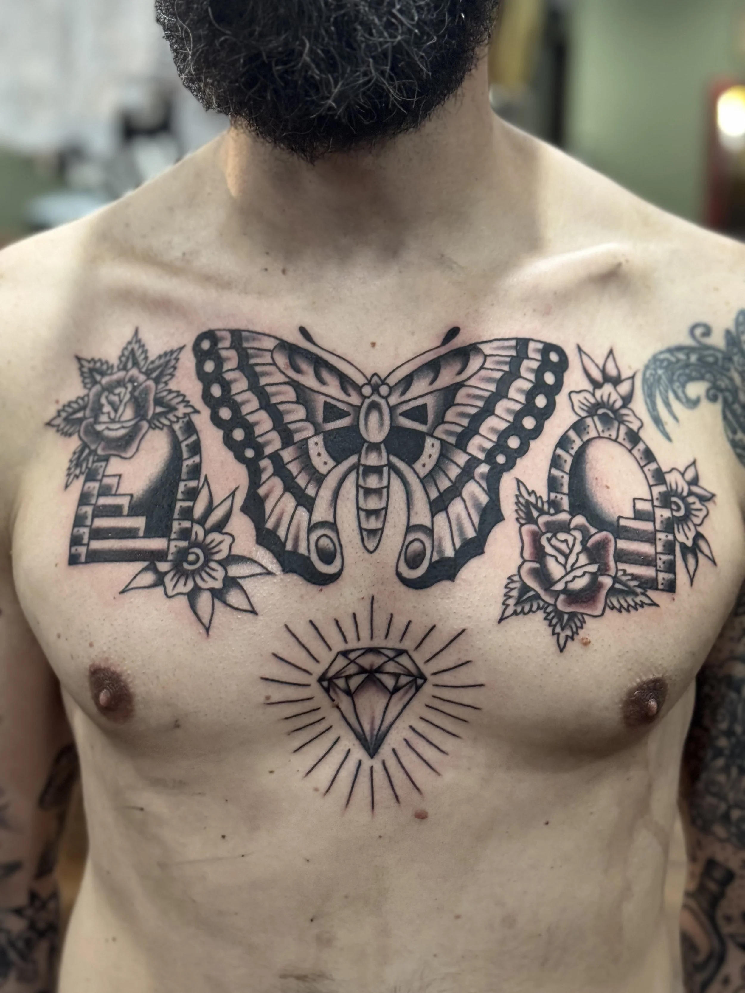 A person with a full beard and tattoos on chest, featuring a large butterfly, a diamond, and roses.