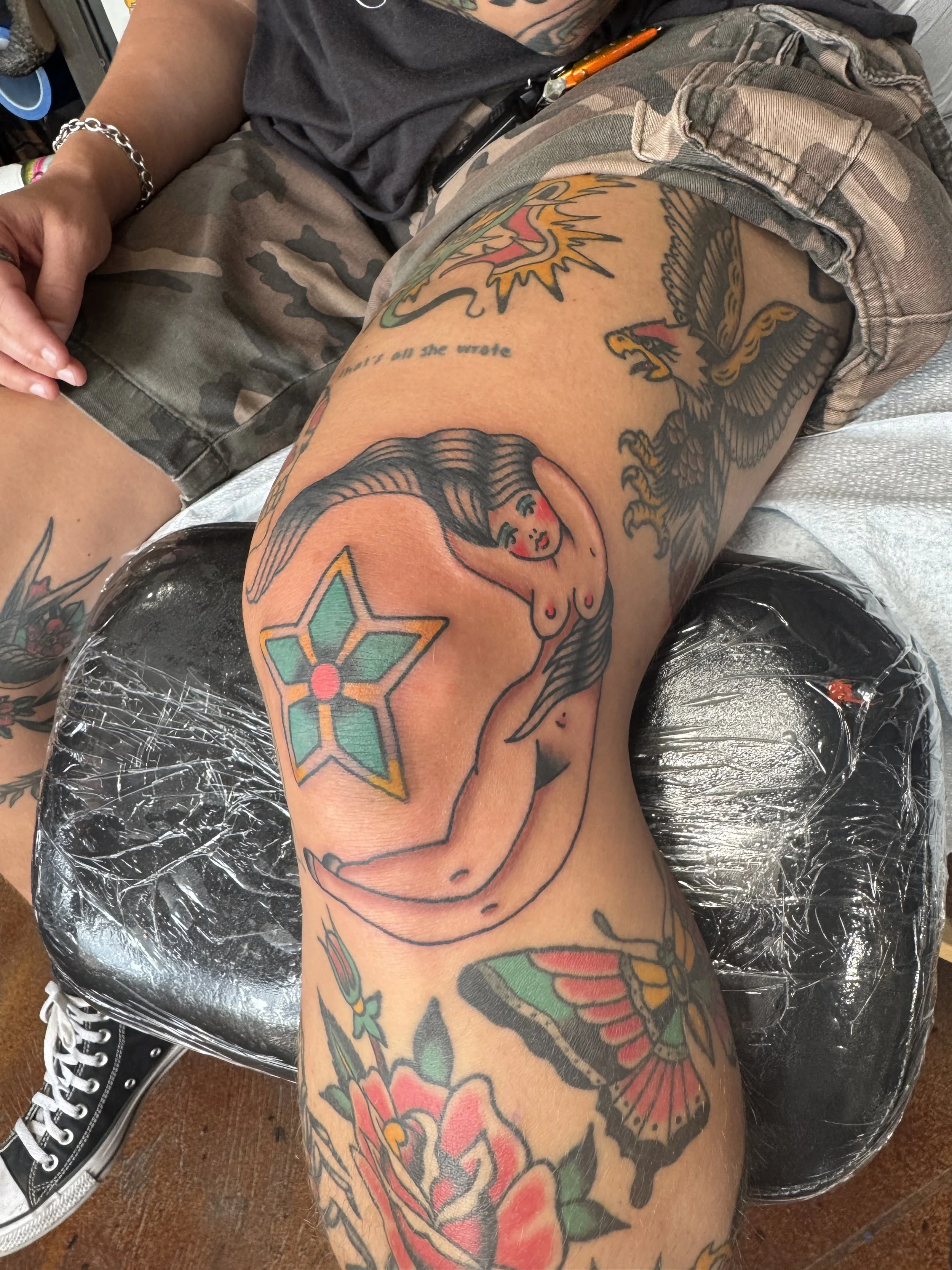 A tattooed person's thigh and knee with various colorful tattoos, including a woman, a snake, a star, a bird, and flowers, sitting on a brown wooden floor with black sneakers and clear plastic wrap covering part of the leg.