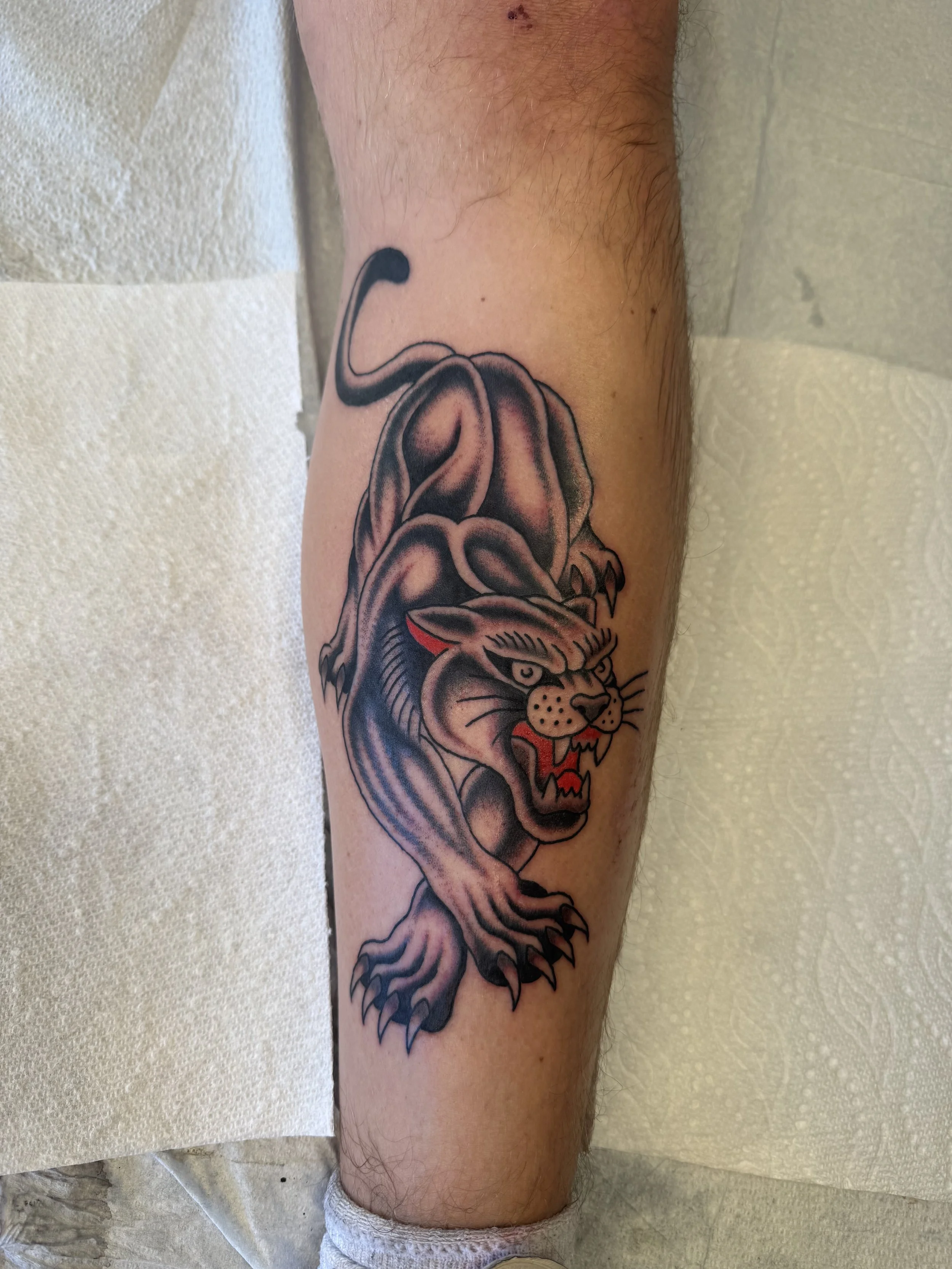 Tattoo of a fierce lion's head on a person's lower leg, with detailed black and gray shading and some red highlights in the mouth.