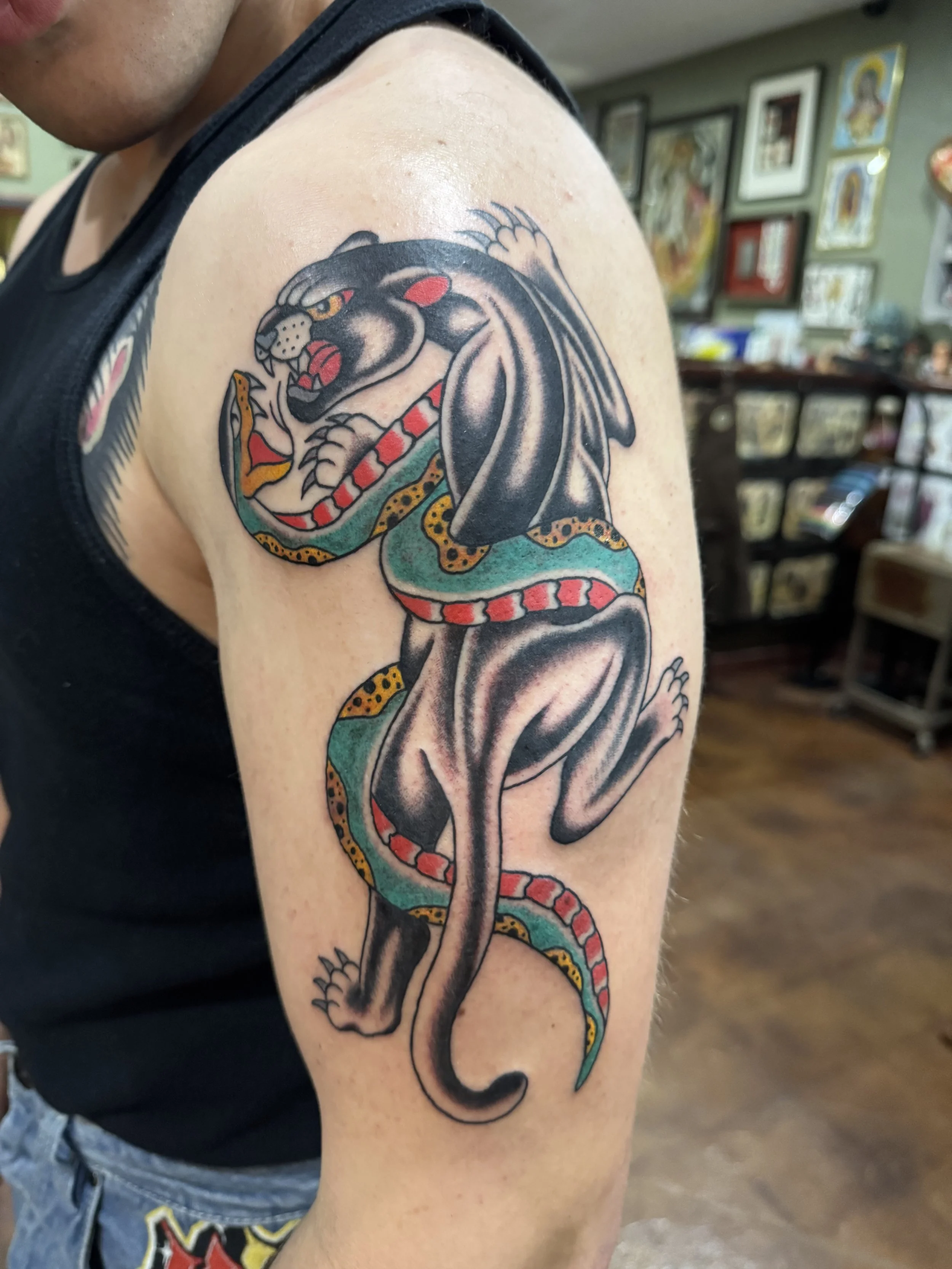 Tattoo of a fierce black panther with yellow eyes and an open mouth, revealing sharp teeth, on a person's upper arm. The panther's body features gray shading and a colorful, swirling design around it.