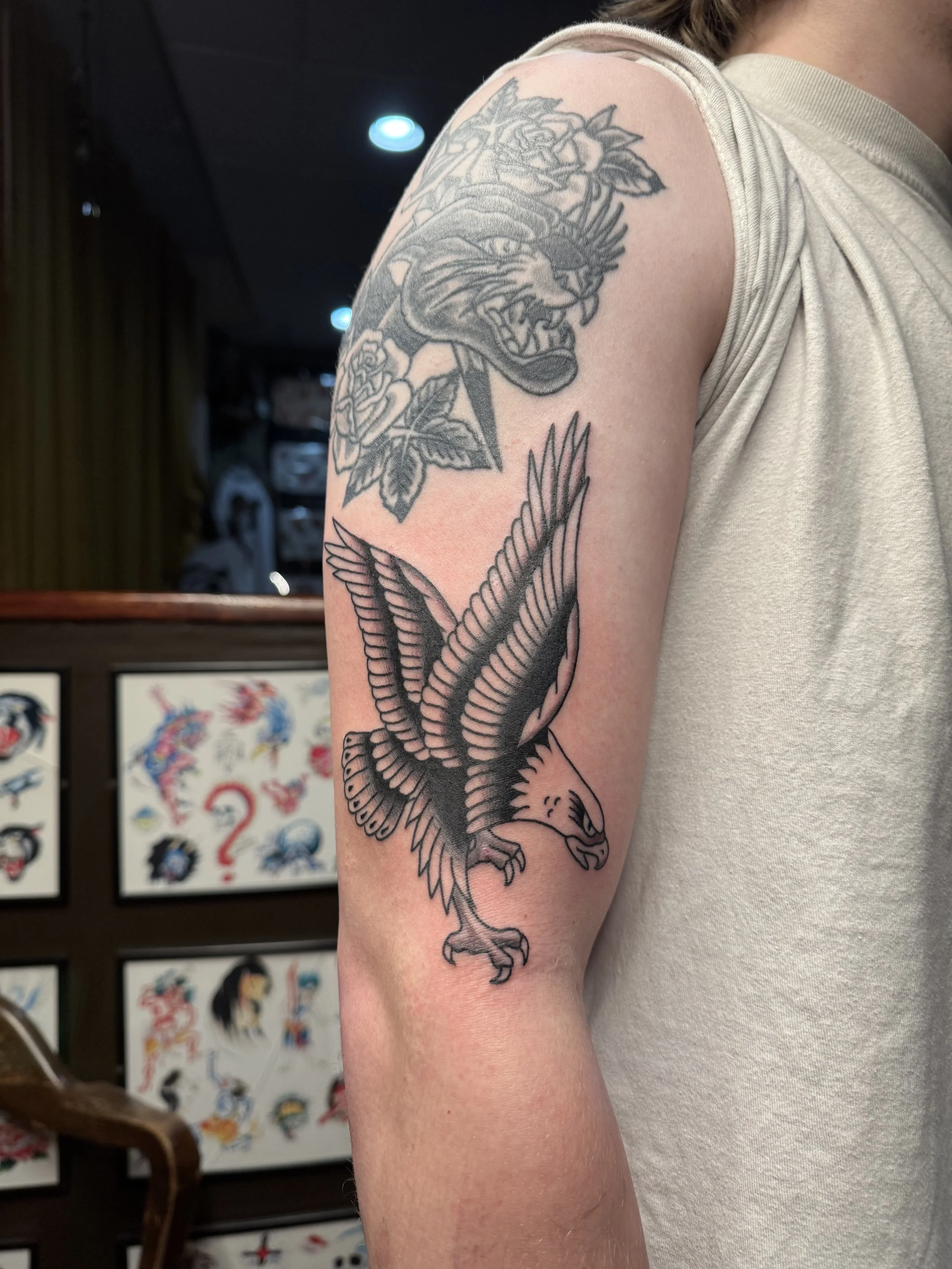 Tattoo of a fierce animal, possibly a jaguar or panther, with roses near its head and a bird with spread wings on the lower arm, on a person's upper arm and shoulder.