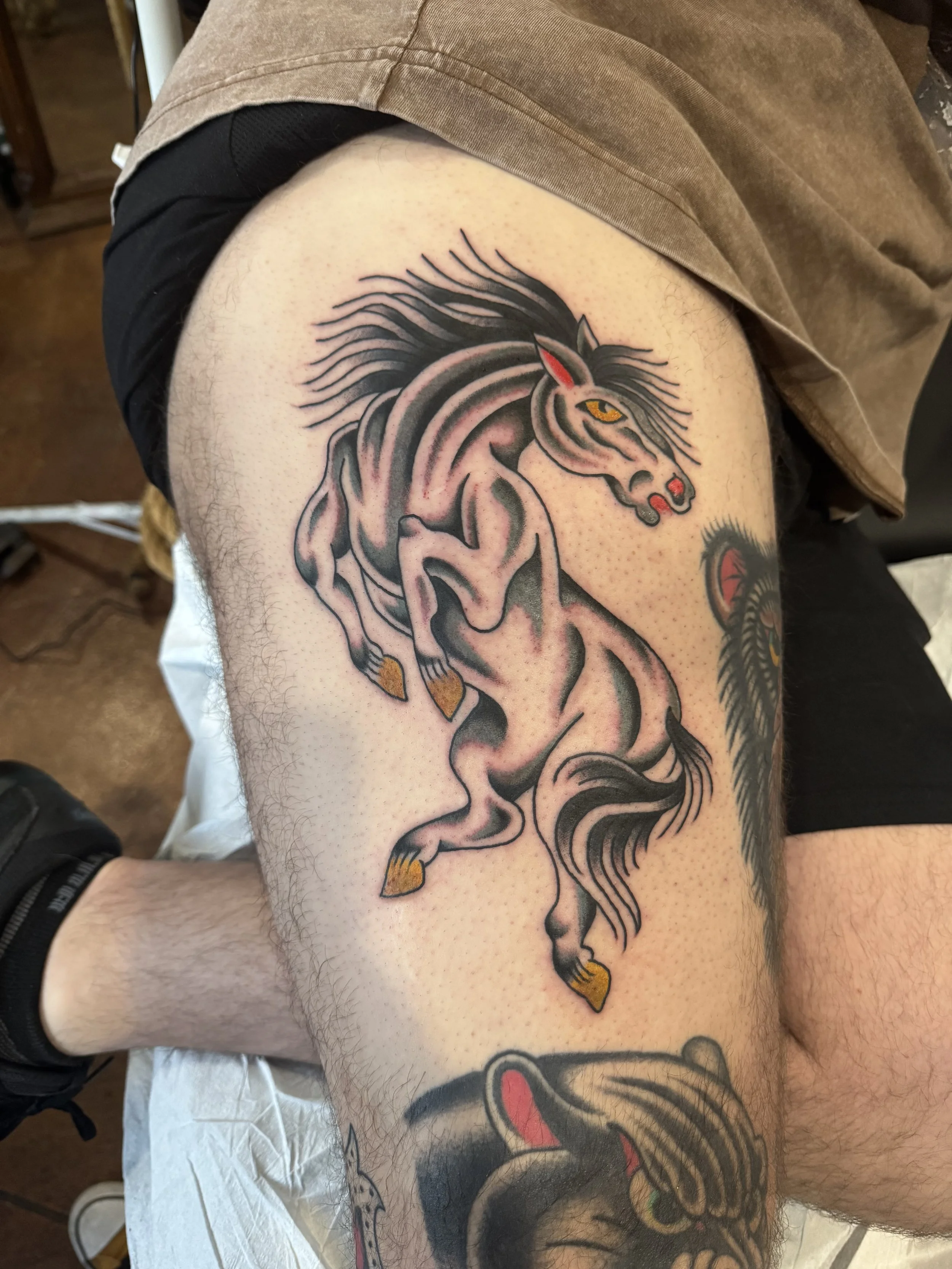 Close-up of a person's thigh with a colorful tattoo of a stylized horse with flowing mane and tail, adorned with yellow and red accents.