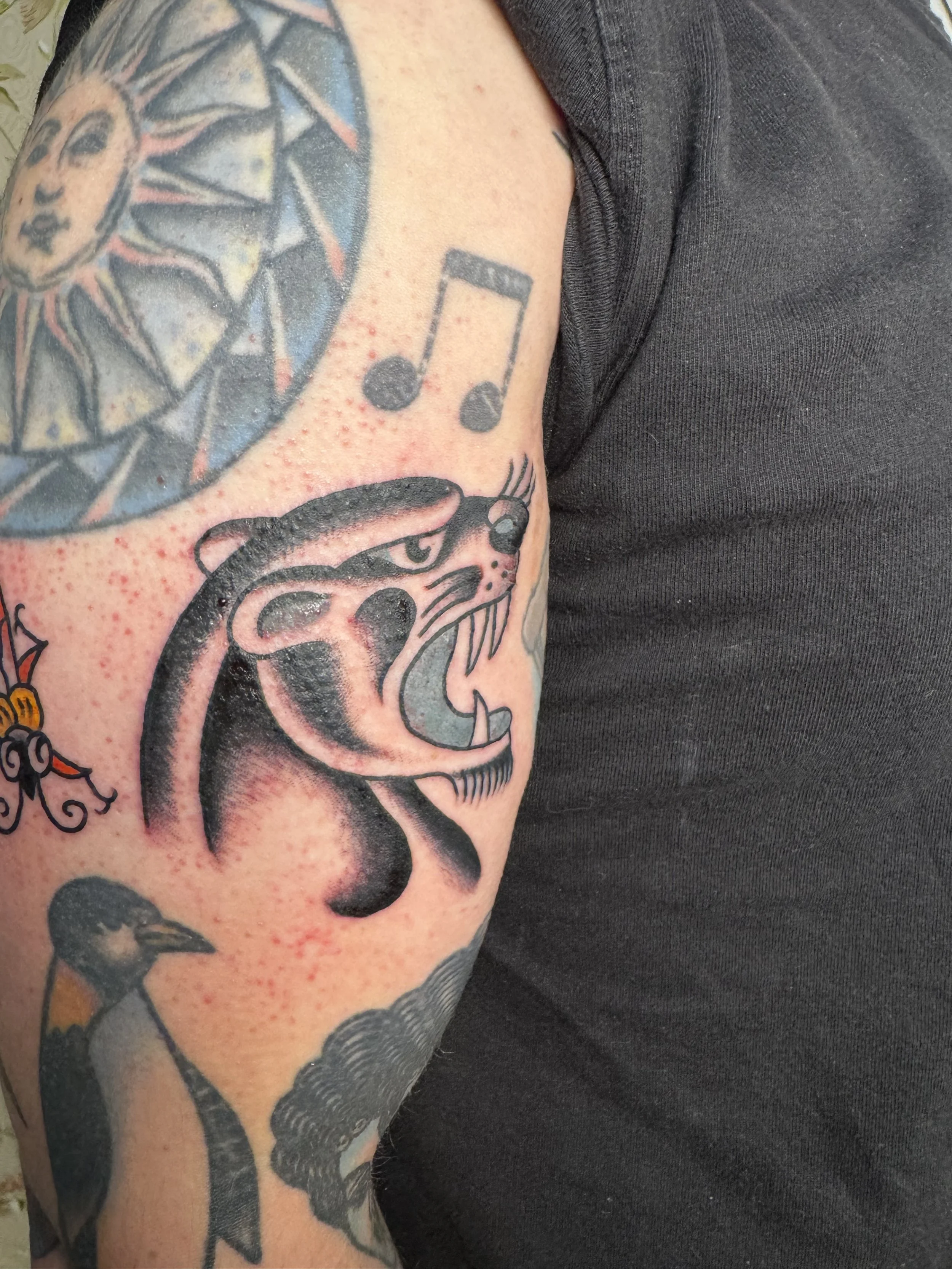 Close-up of a tattooed person's upper arm. The tattoos include a detailed black and gray animal face, musical notes, and a geometric design with skulls and stars.