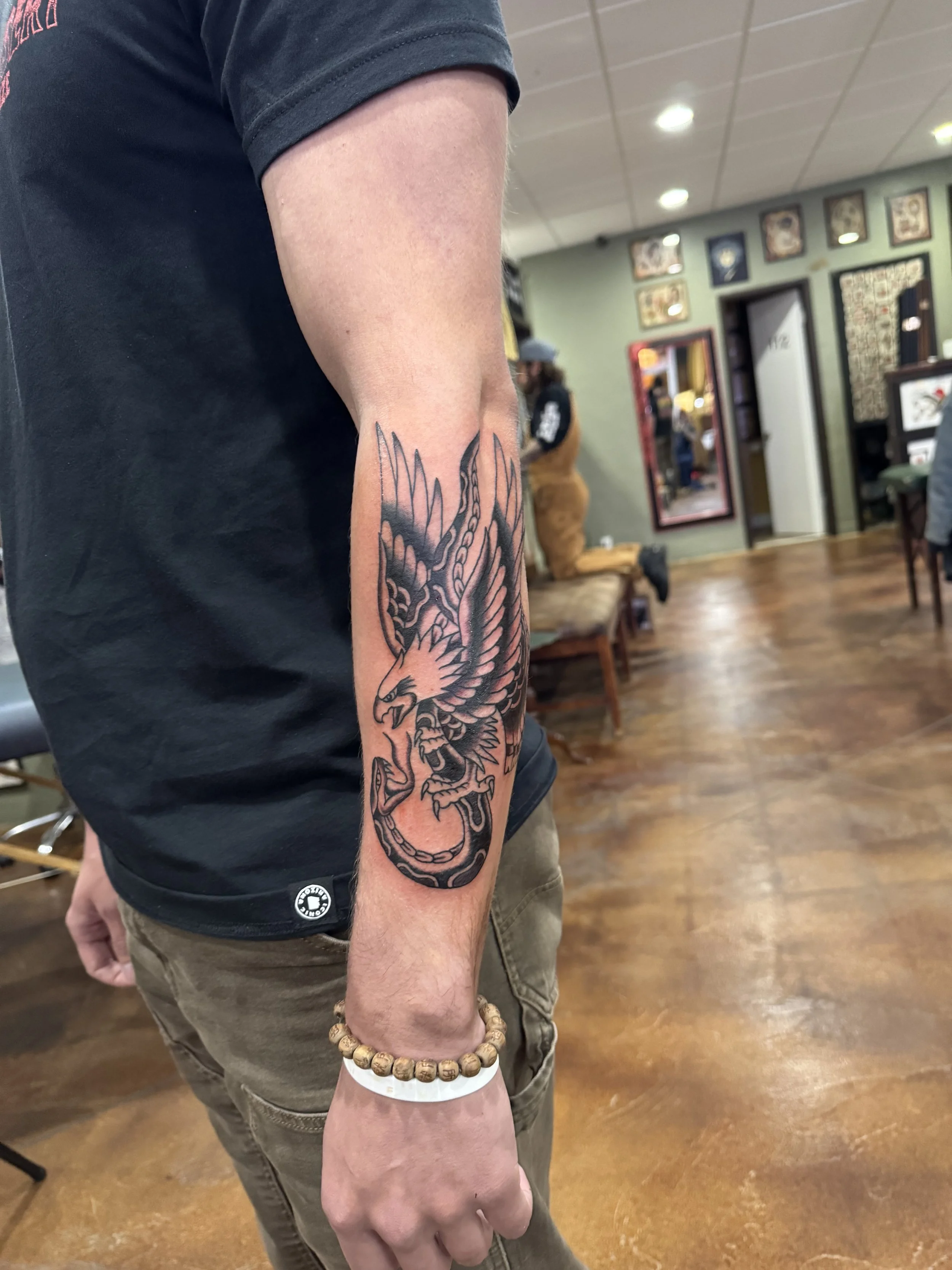 A person's arm with a detailed black and gray tattoo of a fierce bird, possibly an eagle, with its beak open and wings spread. The person is wearing a black t-shirt, a beaded bracelet, and tan pants. The background shows a tattoo studio with wooden f