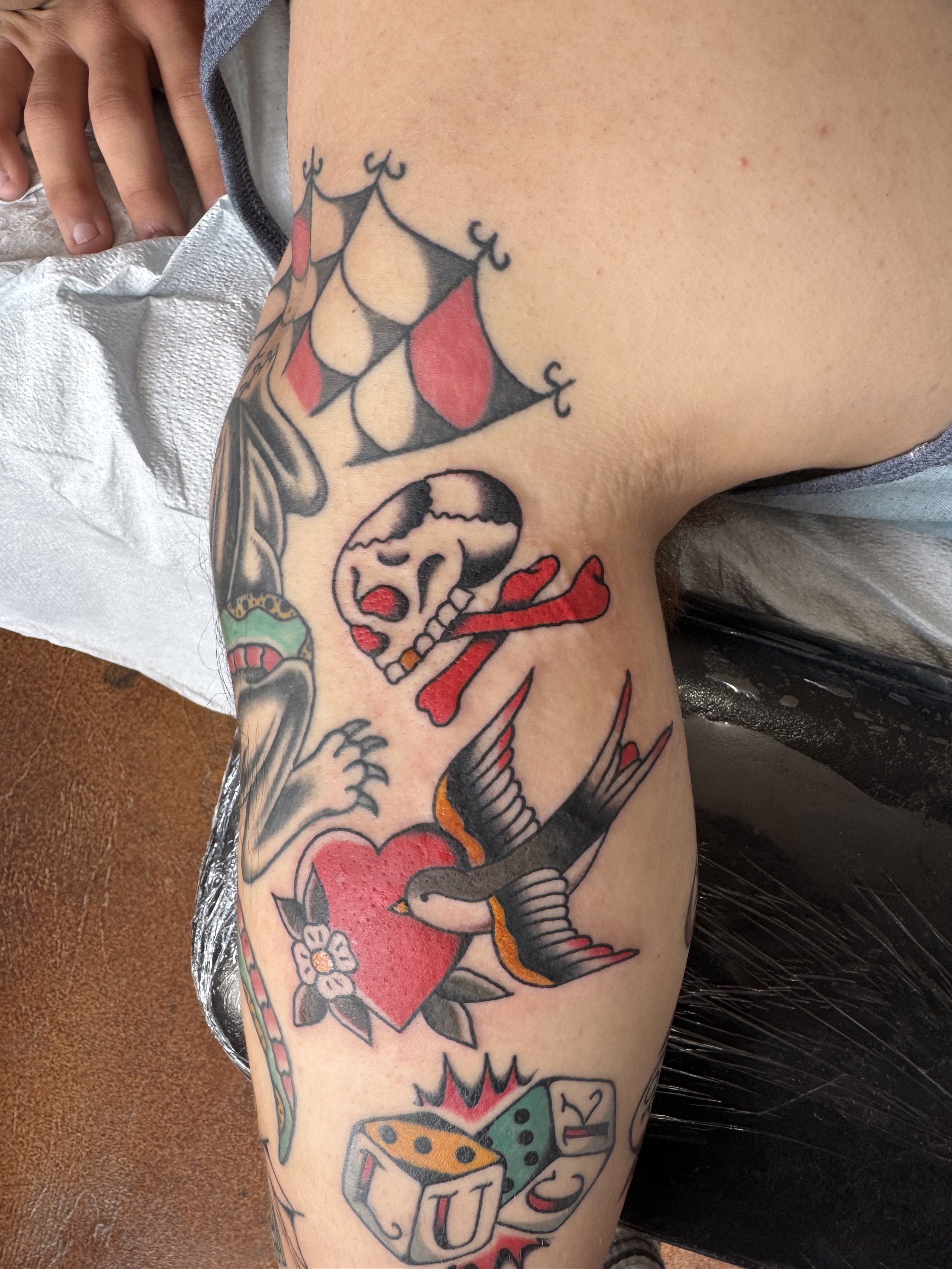 Colorful tattoo featuring a skull with a rose, a large heart, a fish, a dragonfly, and various decorative elements on a person's thigh.
