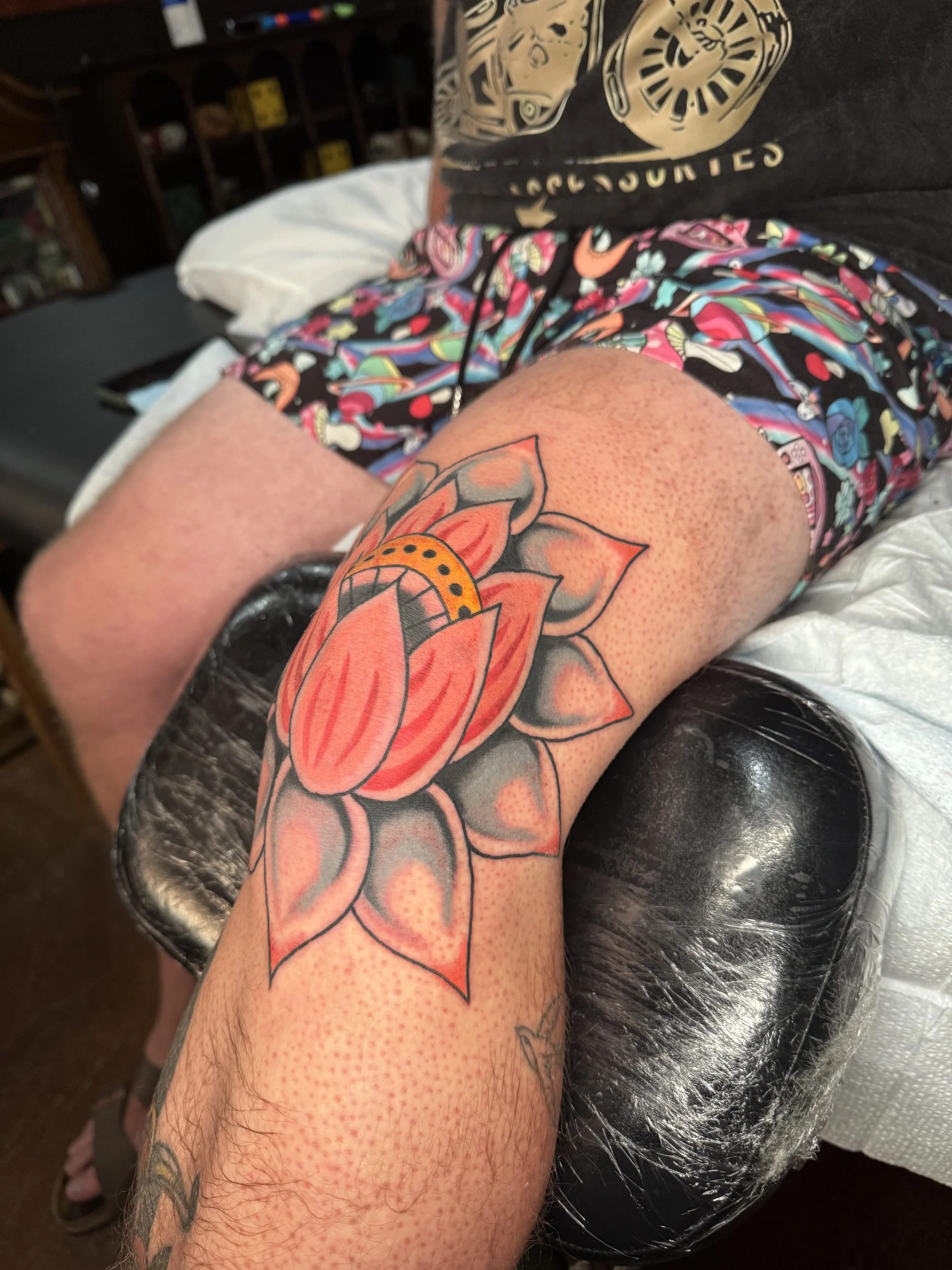 A person's arm resting on a black padded armrest with a large colorful tattoo of a flower. The person is wearing colorful shorts with a pattern and a black t-shirt with a gold design. The setting appears to be a tattoo or tattooing environment.