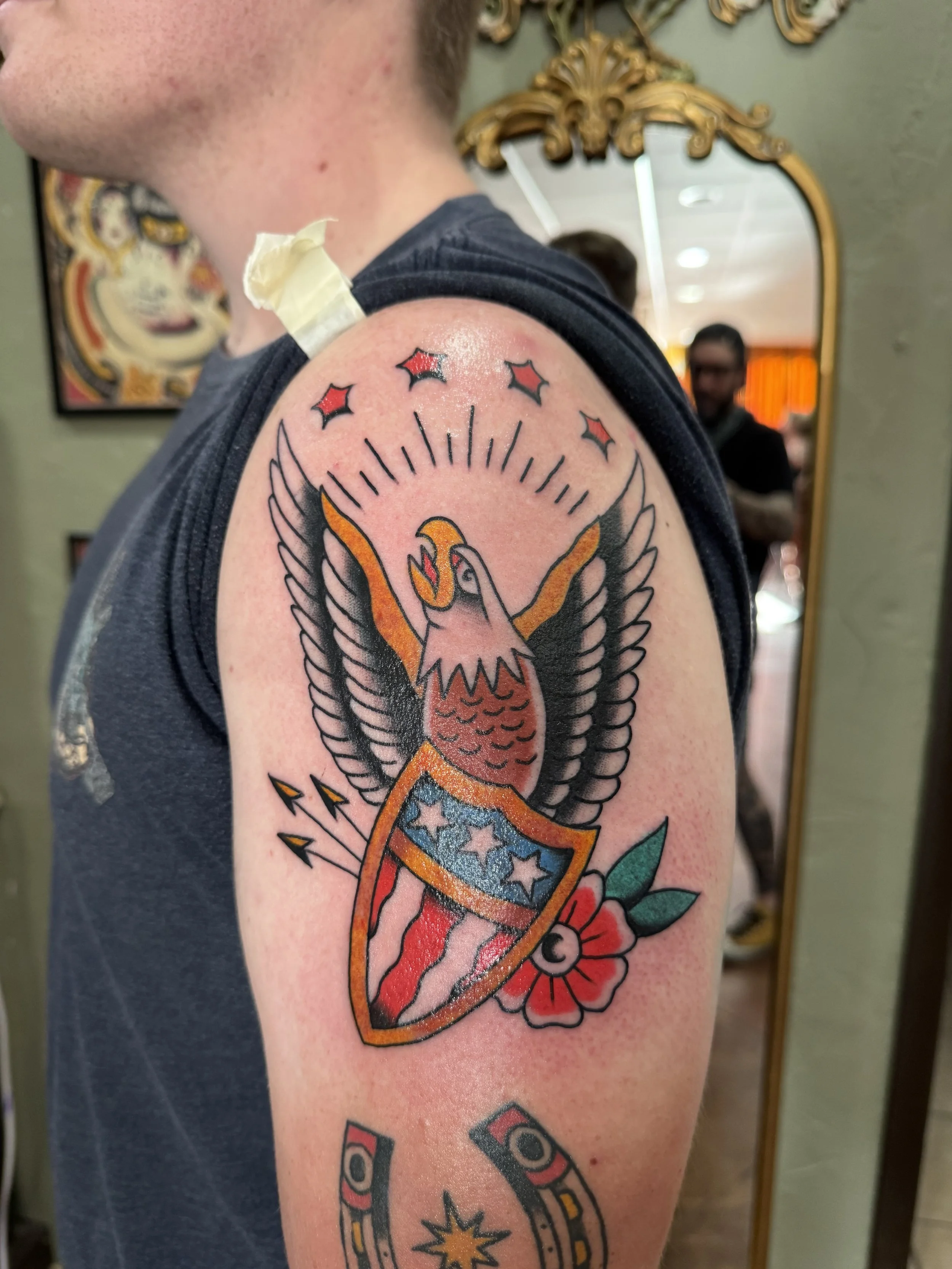 Close-up of a person's upper arm with a tattoo of a phoenix with spread wings and stars, and a shield with stars. The person is wearing a dark sleeveless shirt, and there is medical tape on the shoulder. The background includes a mirror and framed ar