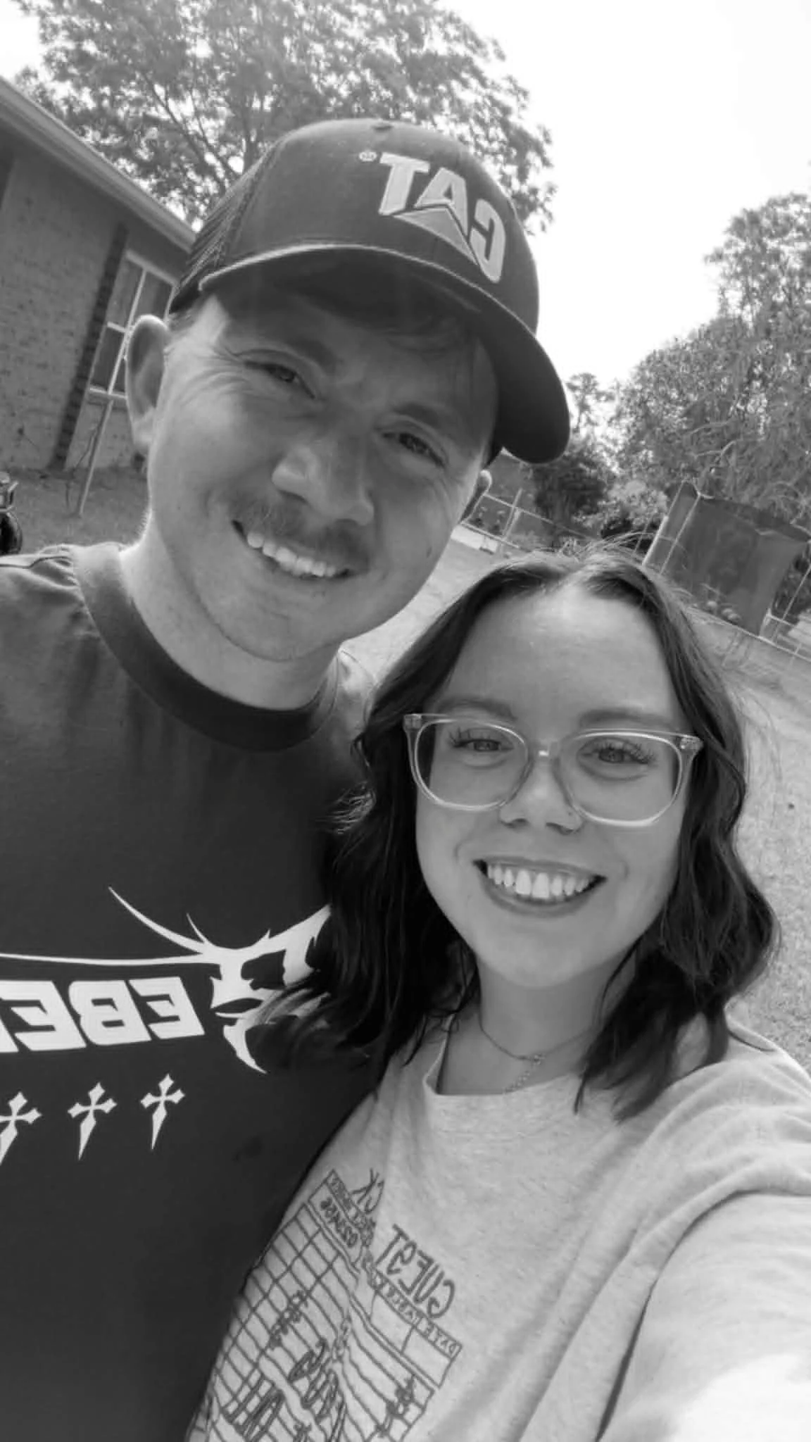 A black and white photo of a young man and woman smiling outdoors, with trees and a house in the background.