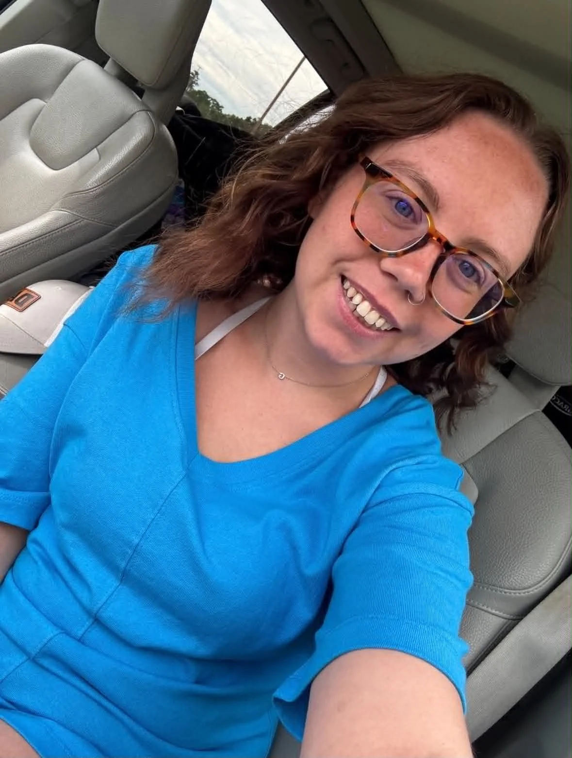 A smiling woman with glasses and a septum piercing sitting in a car, wearing a blue shirt, in a selfie pose.