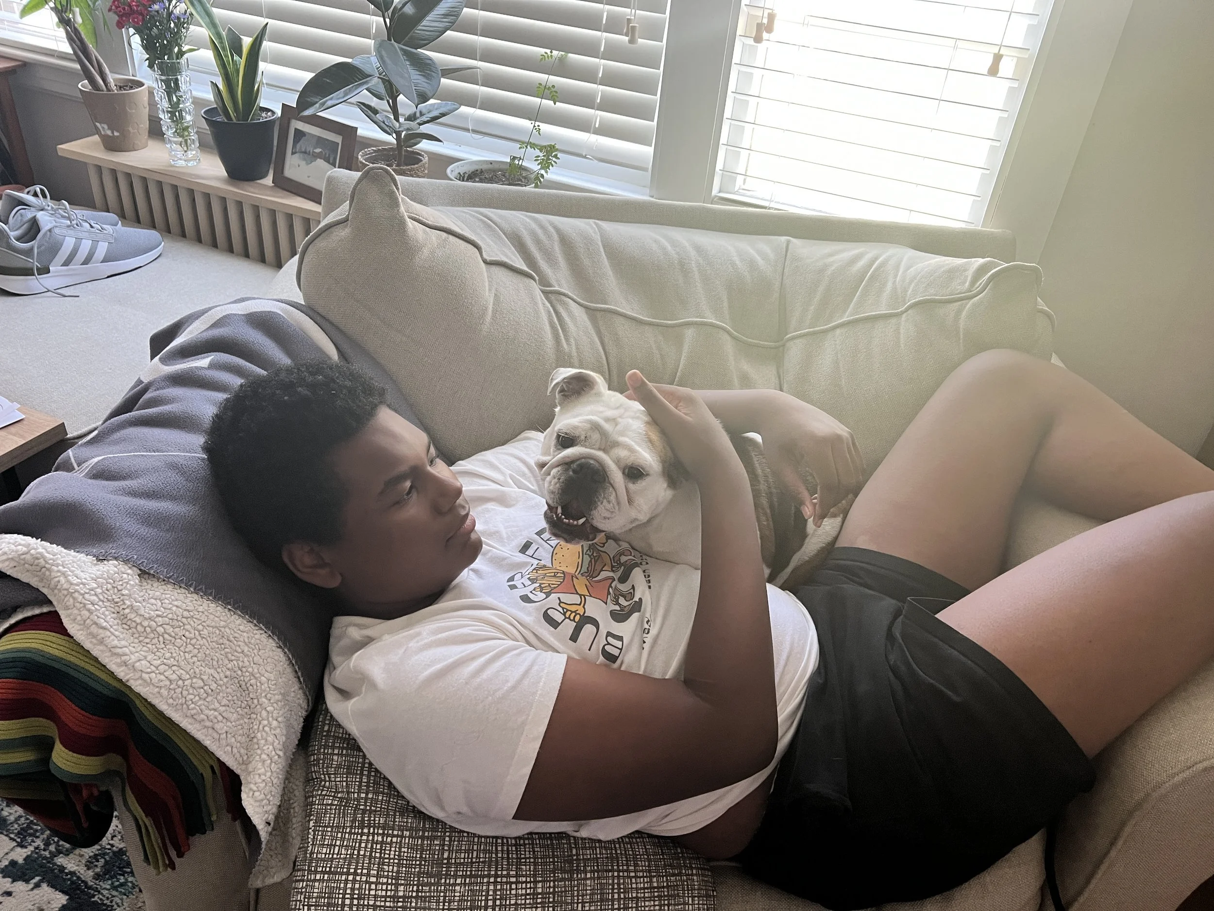 A young man with short dark hair is lying on a couch, cuddling a white bulldog with a brown patch over one eye, in a living room with sunlight coming through white blinds. There are plants, a picture frame, and a pair of sneakers on the windowsill be
