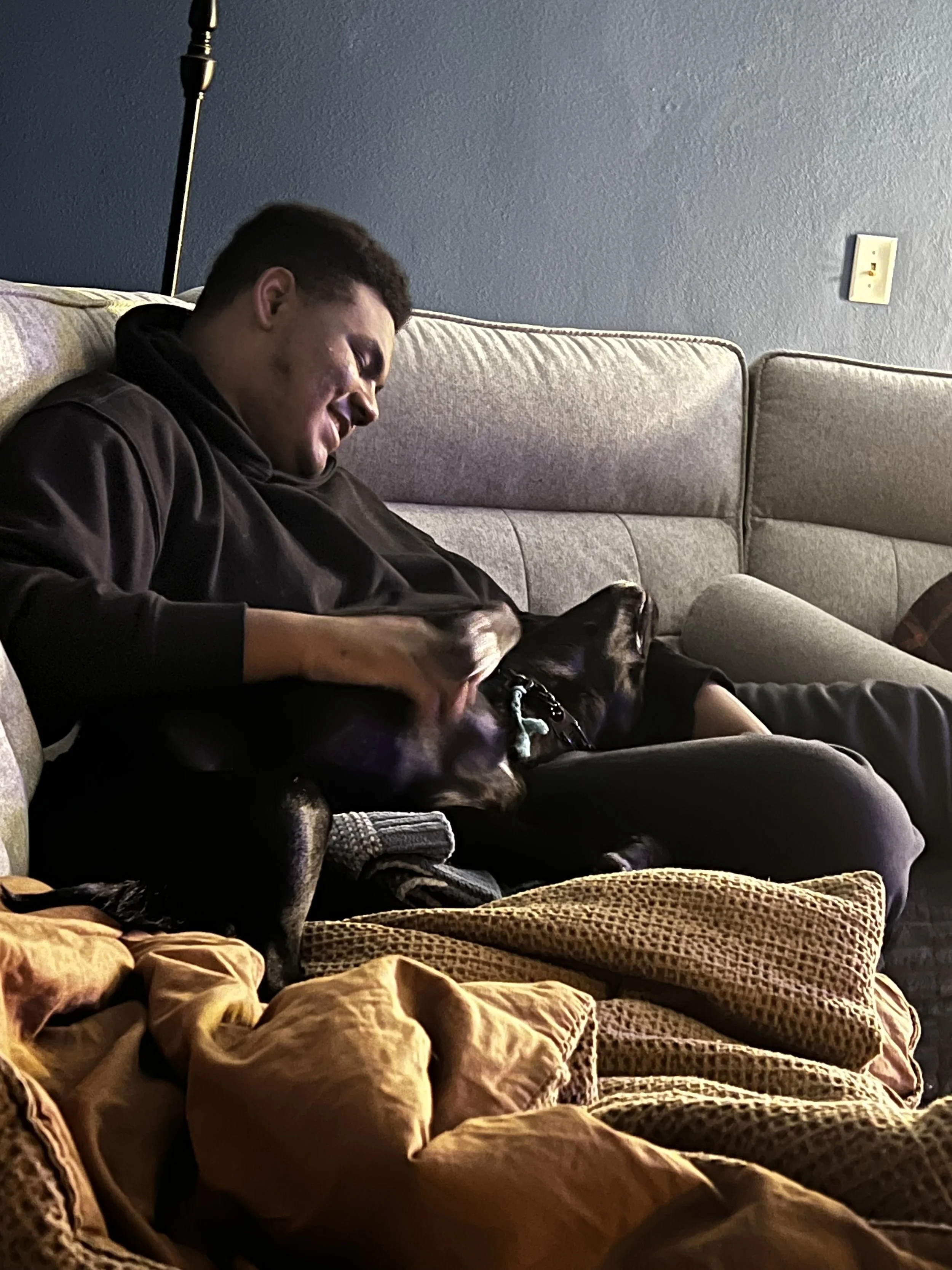 A young man with short dark hair and a mole on his face, smiling and sitting on a beige couch, petting a large black dog with a green collar. There is a pile of yellow and patterned blankets or clothing in the foreground, and a blue wall with a black