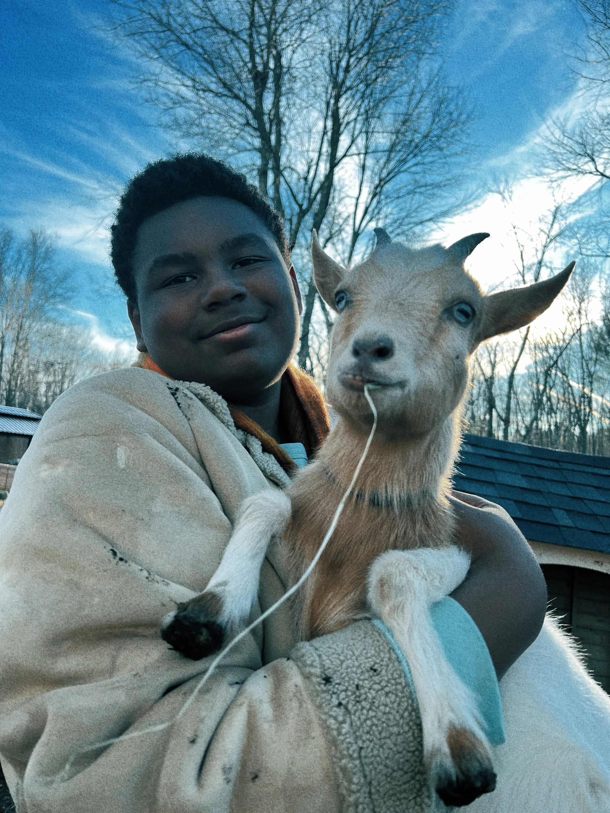 A person with dark skin and short curly hair holding a light-colored goat outdoors during daylight with a background of leafless trees and a blue sky.