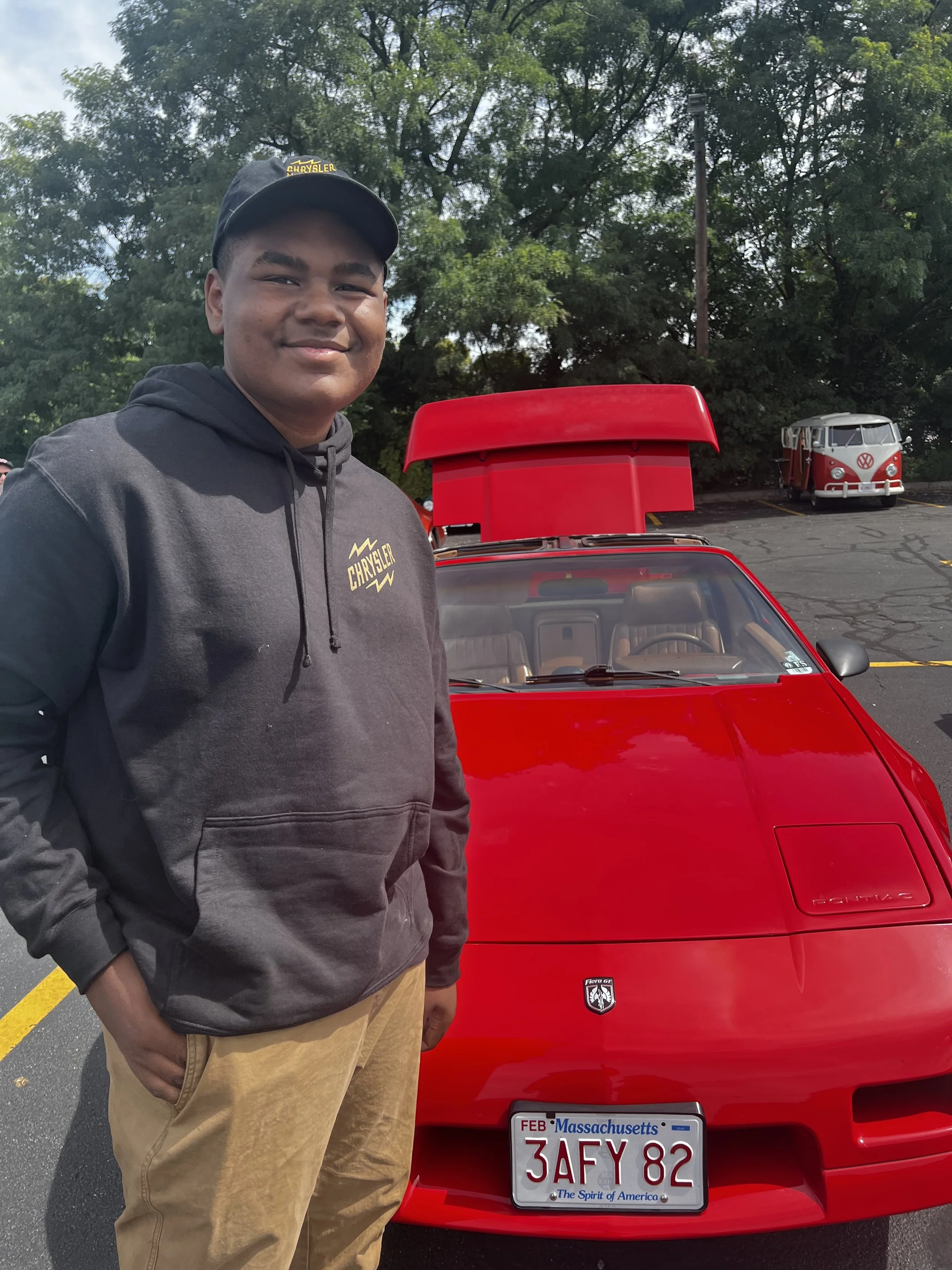 A young man wearing a black hoodie and a black cap with yellow text stands next to a red sports car with a Massachusetts license plate. The car has a small emblem on the front and its hood is partially open. In the background, there is a parking lot with a Volkswagen Type 2 van with a red and white paint scheme, surrounded by trees.