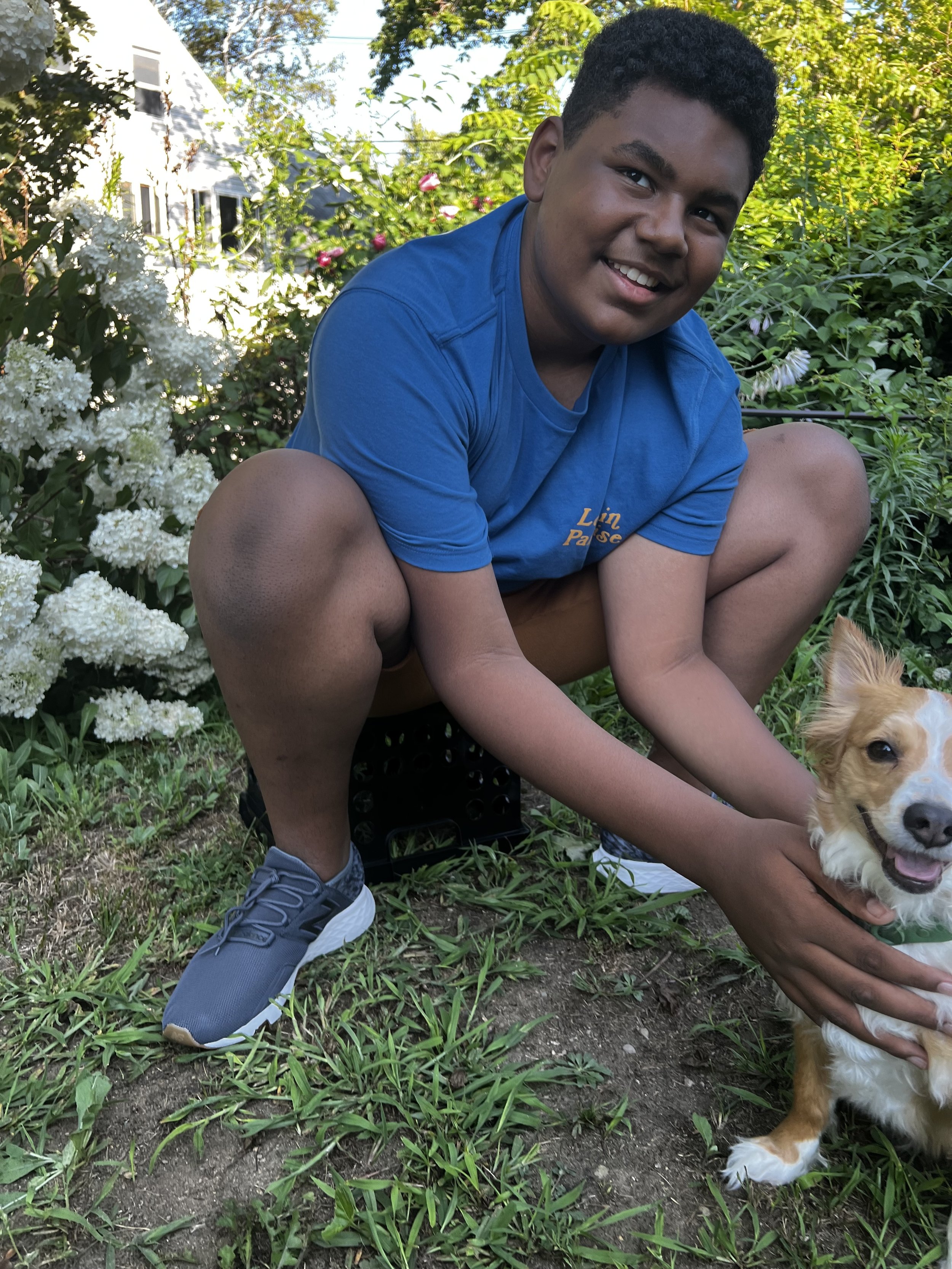 A young boy with dark curly hair, wearing a blue T-shirt, gray athletic shoes, and shorts, crouching in a garden. He is smiling and holding a small, golden-haired dog with white markings on its face and chest. Flowers and greenery surround them, with