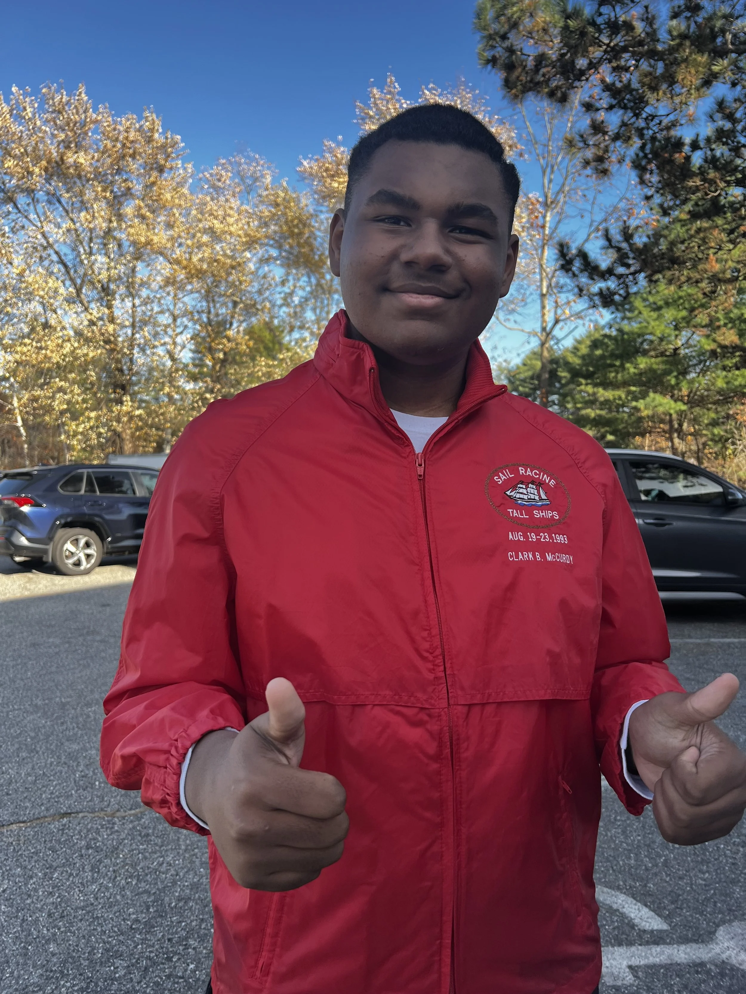 A smiling young man with short hair wearing a red jacket, pointing both thumbs up, standing outdoors in a parking lot with trees and cars in the background on a clear day.
