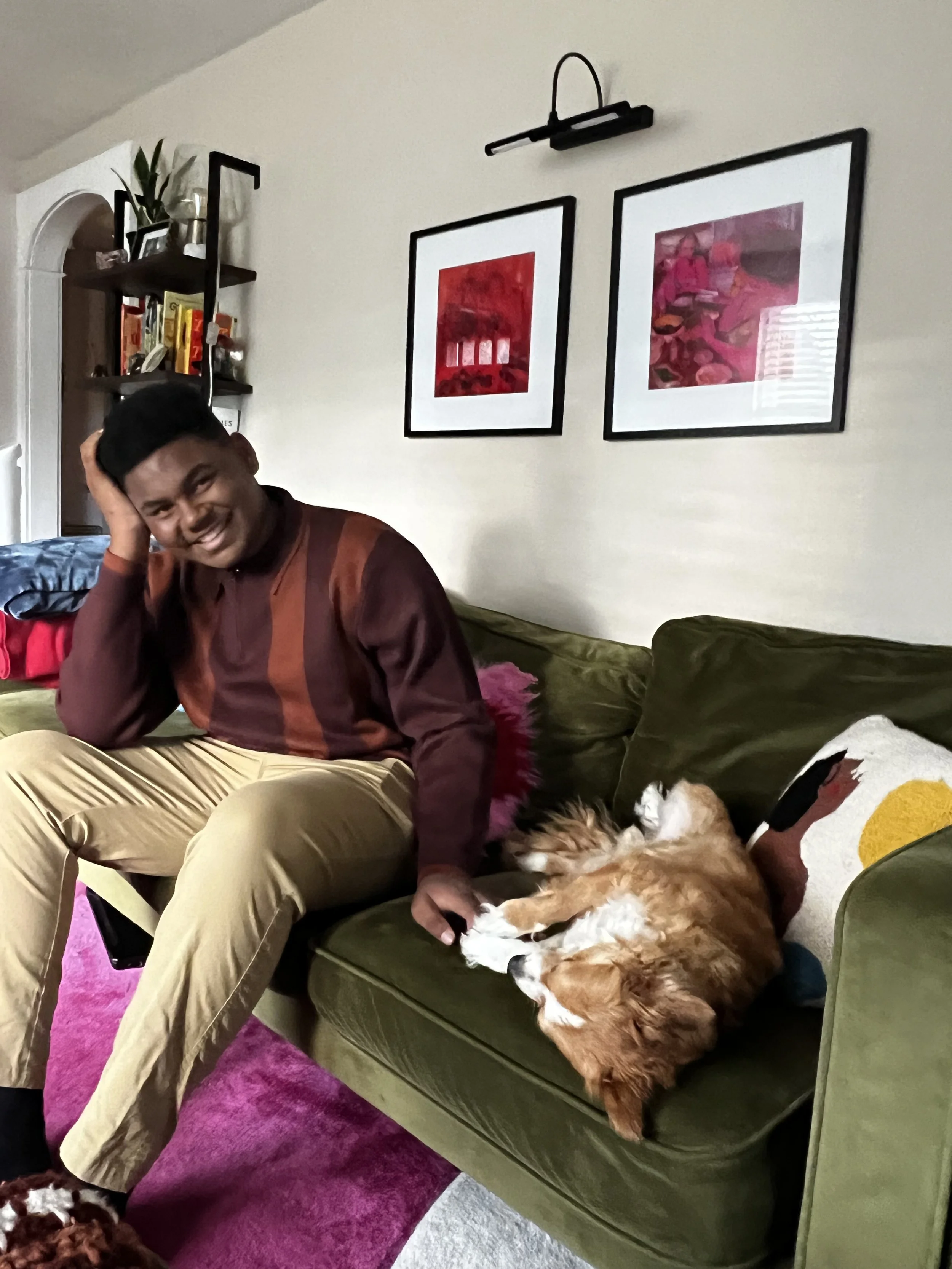 A man with dark hair smiling, sitting on a green sofa with a hand on a relaxed, large brown and white dog lying on its back. The dog is close to the man's lap. Behind them, there are framed pictures on the wall, shelves with books and decor, a couch 